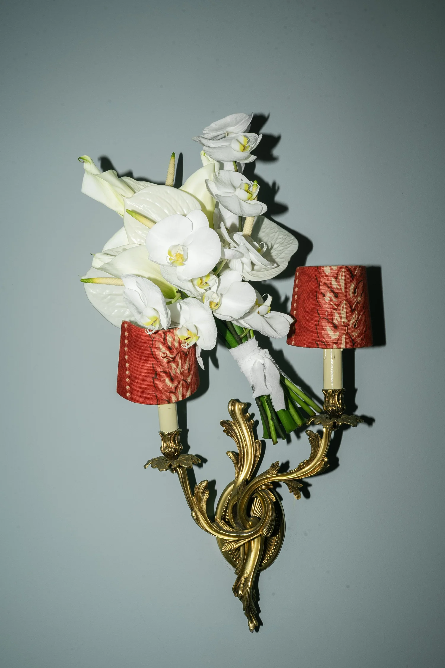White orchids and calla lilies arranged on an ornate gold wall sconce with red lampshades