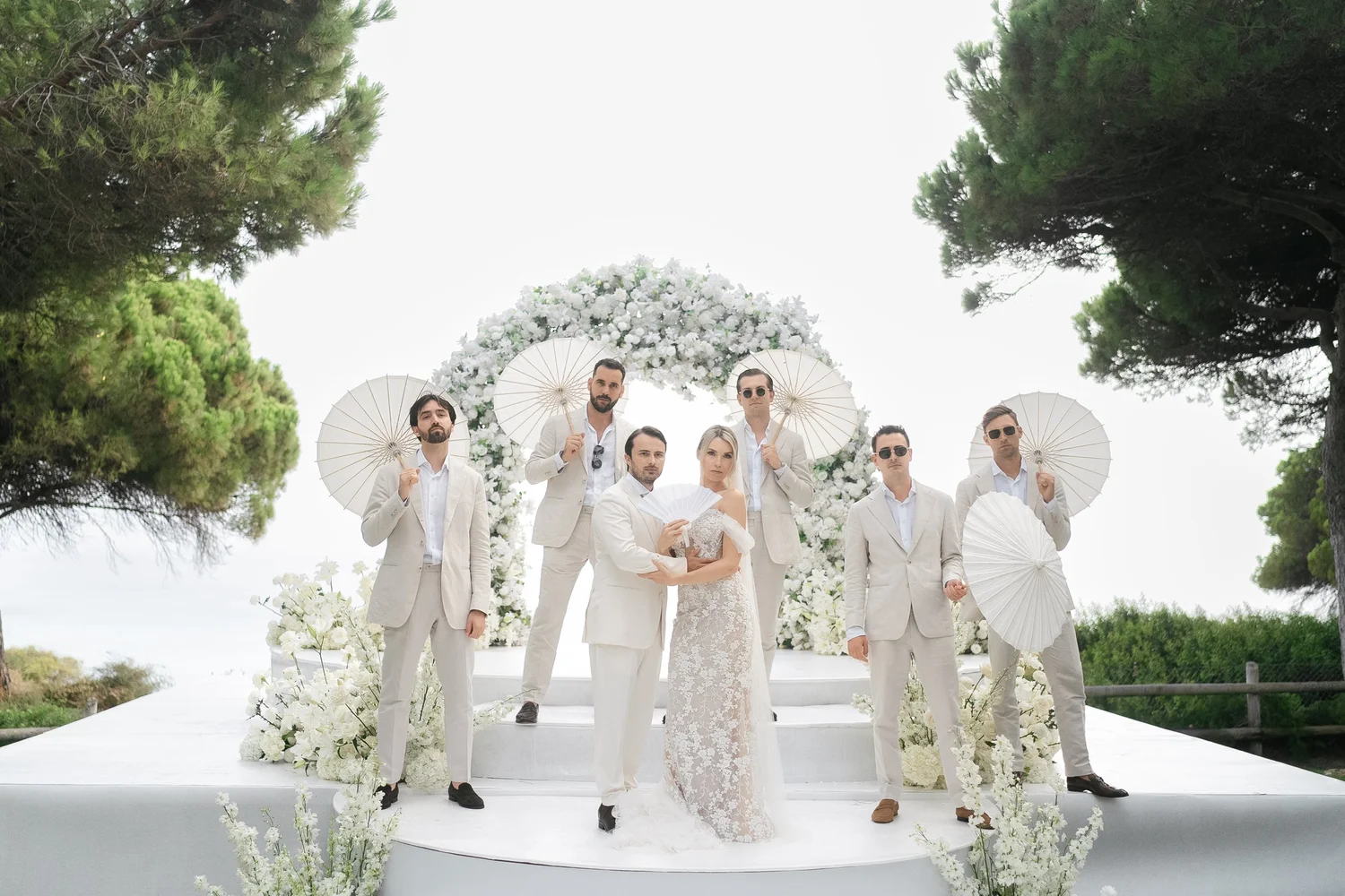 Bride and groom with their wedding party in pale suits beneath a white floral arch