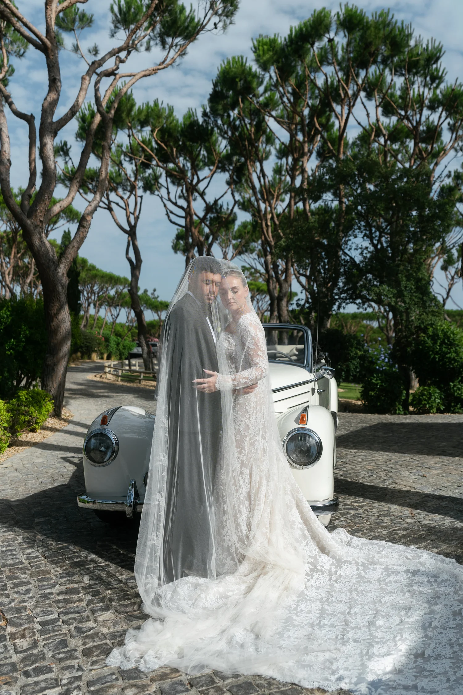 Newlyweds embracing beneath the bride’s veil in front of a white vintage convertible