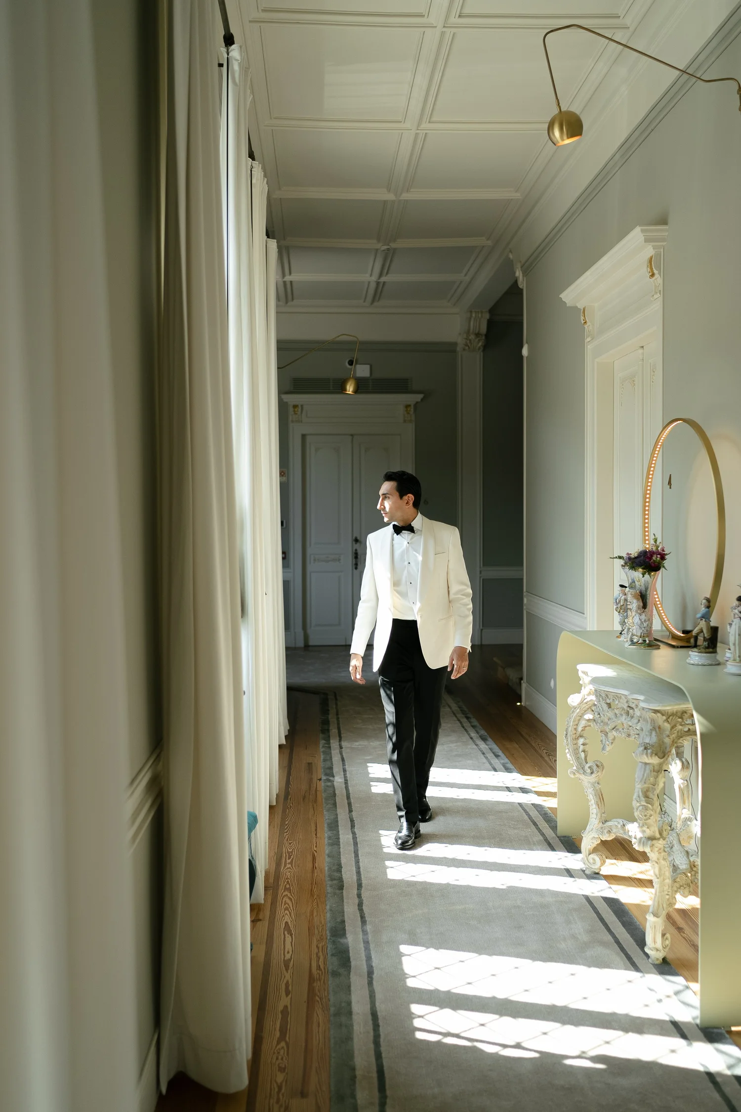Groom in an ivory dinner jacket walking through a sunlit hallway before the wedding