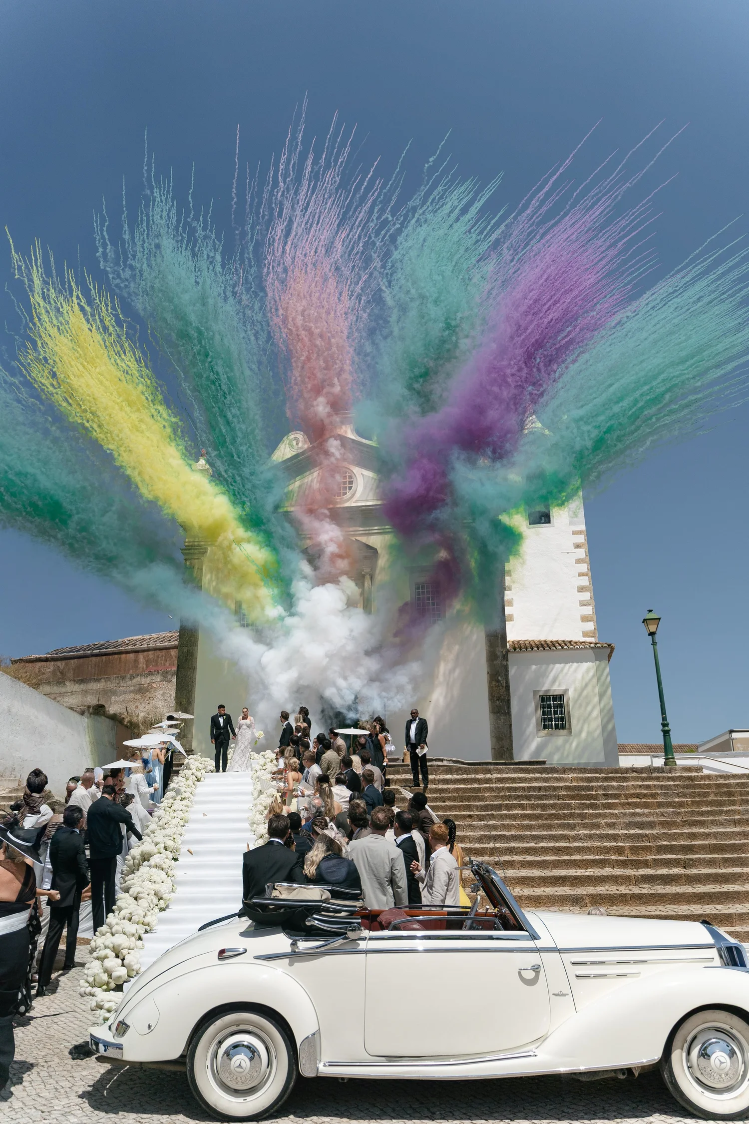 Colourful smoke above a church wedding exit, with guests and a white vintage convertible below