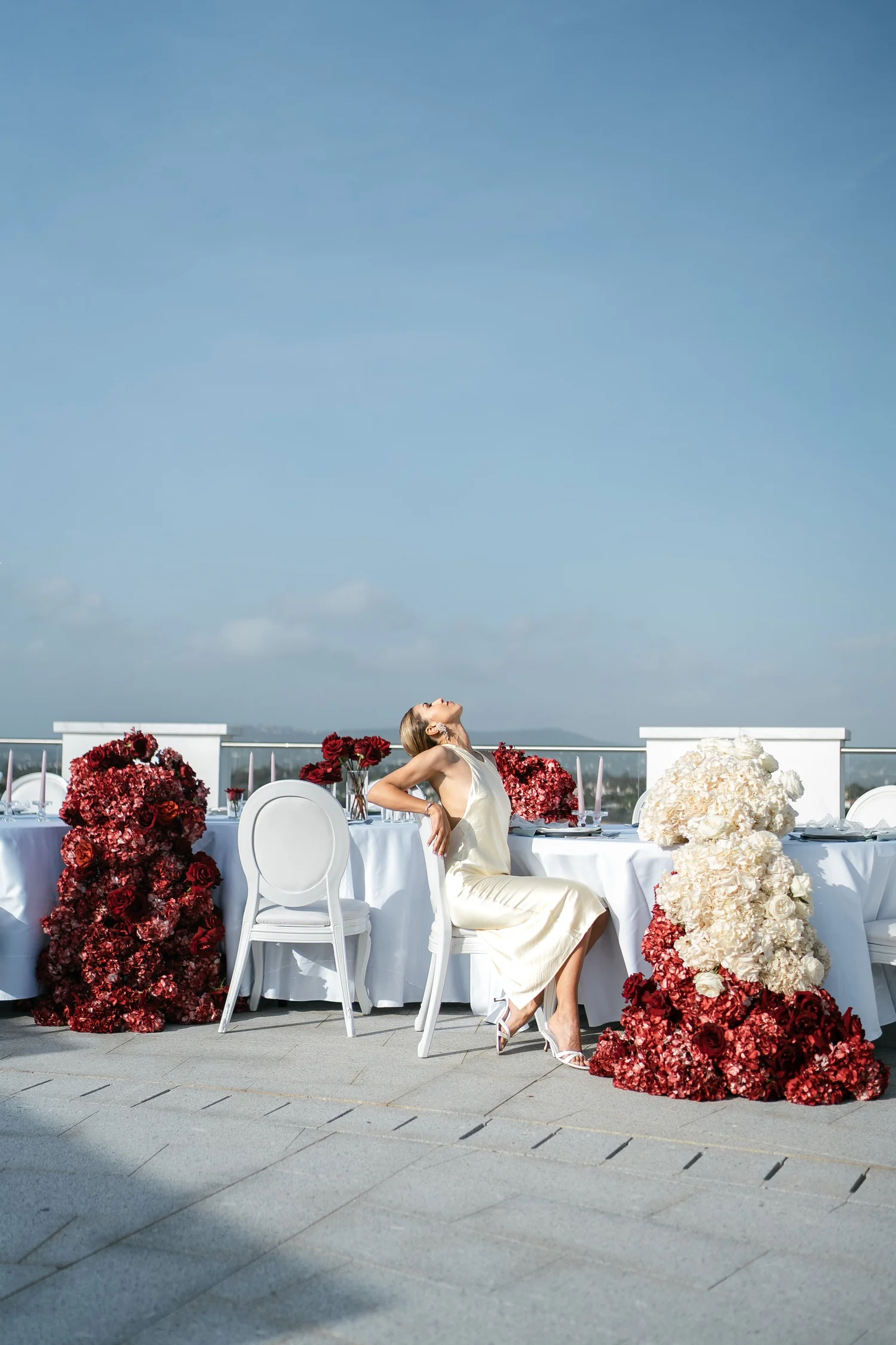 Bride in an ivory satin dress seated on a terrace among red and white wedding flowers