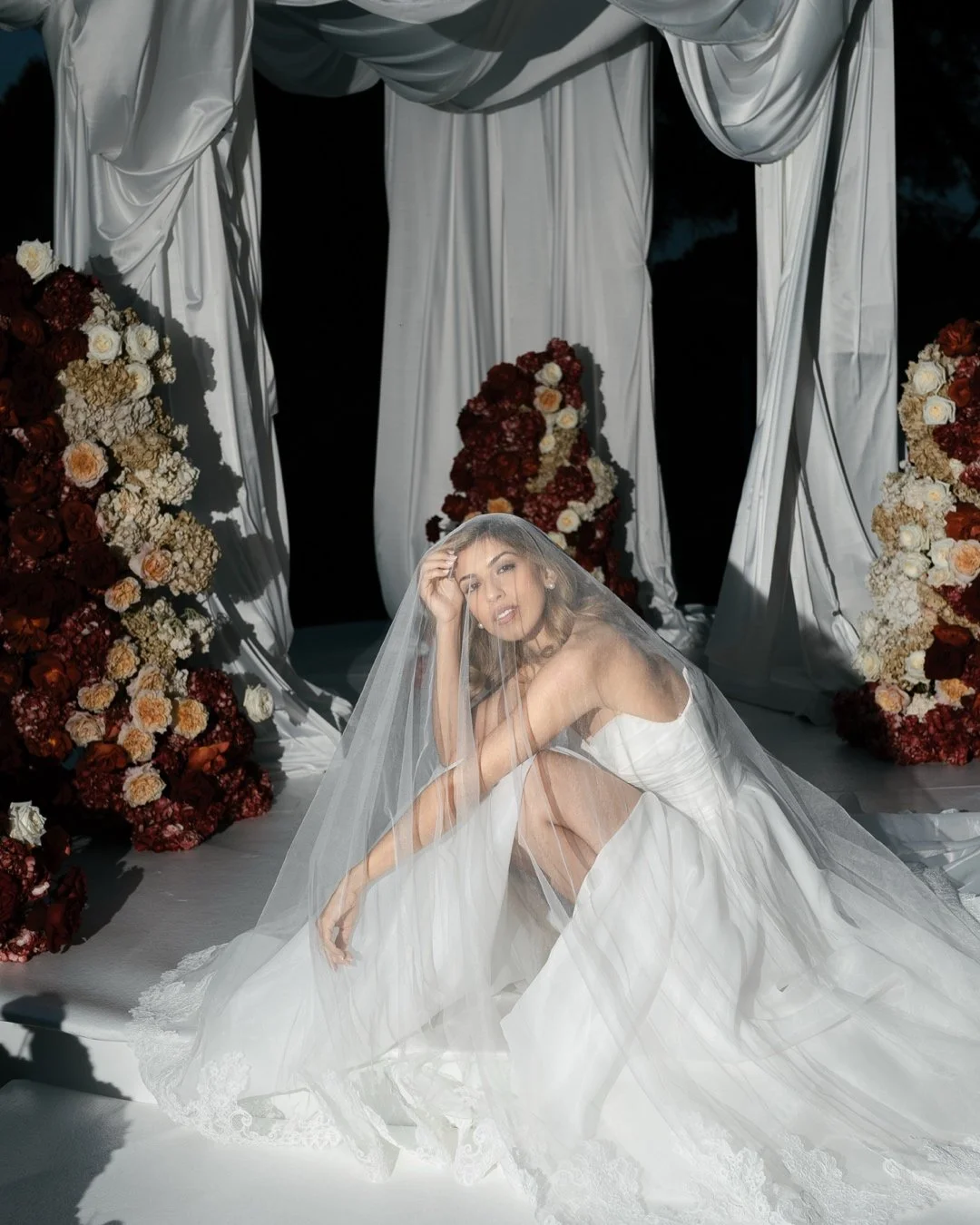 Bride beneath her veil, seated among wedding flowers and white fabric drapes