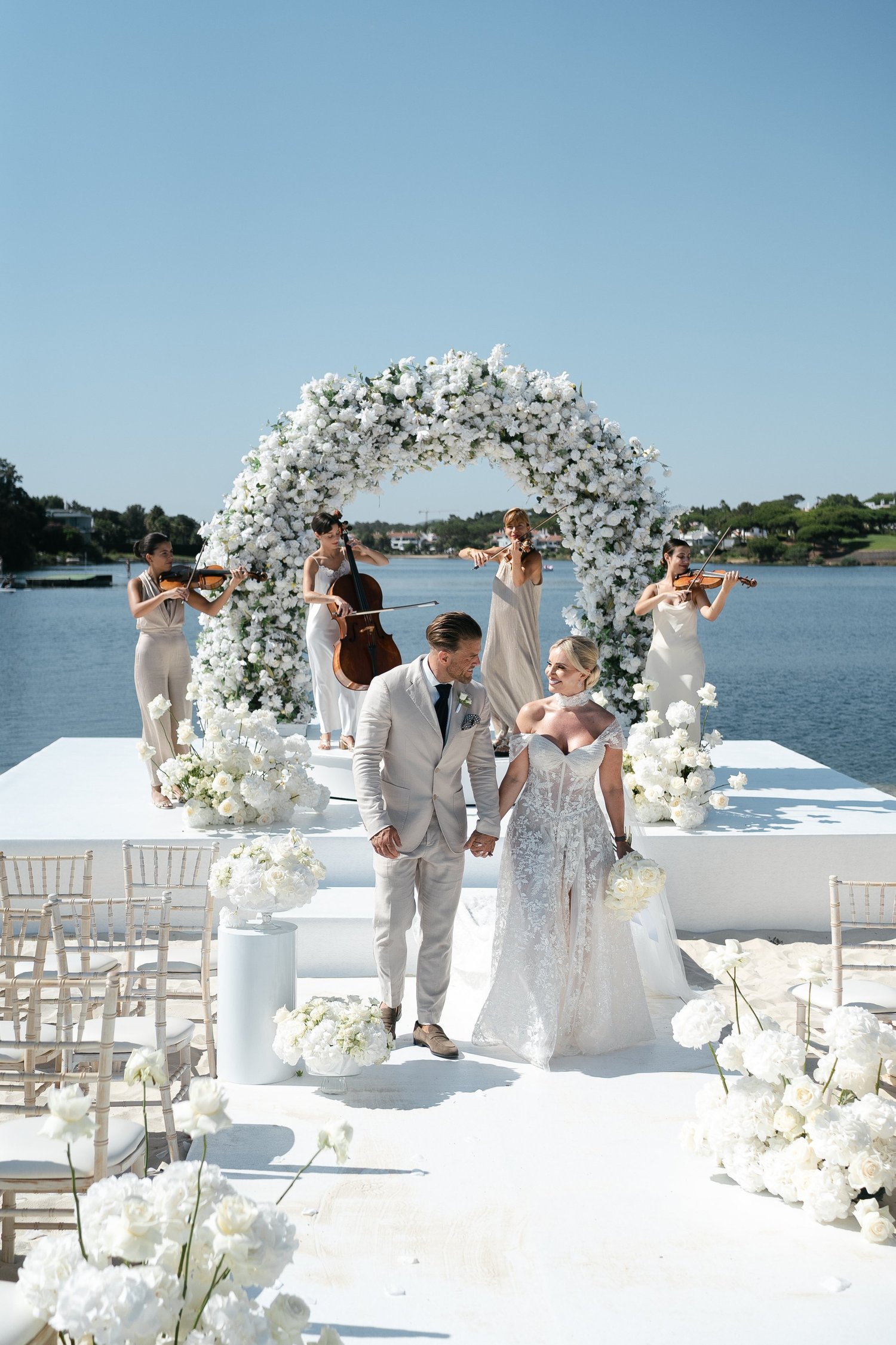 Bride and groom leaving a waterside ceremony with a white floral arch and string musicians behind them