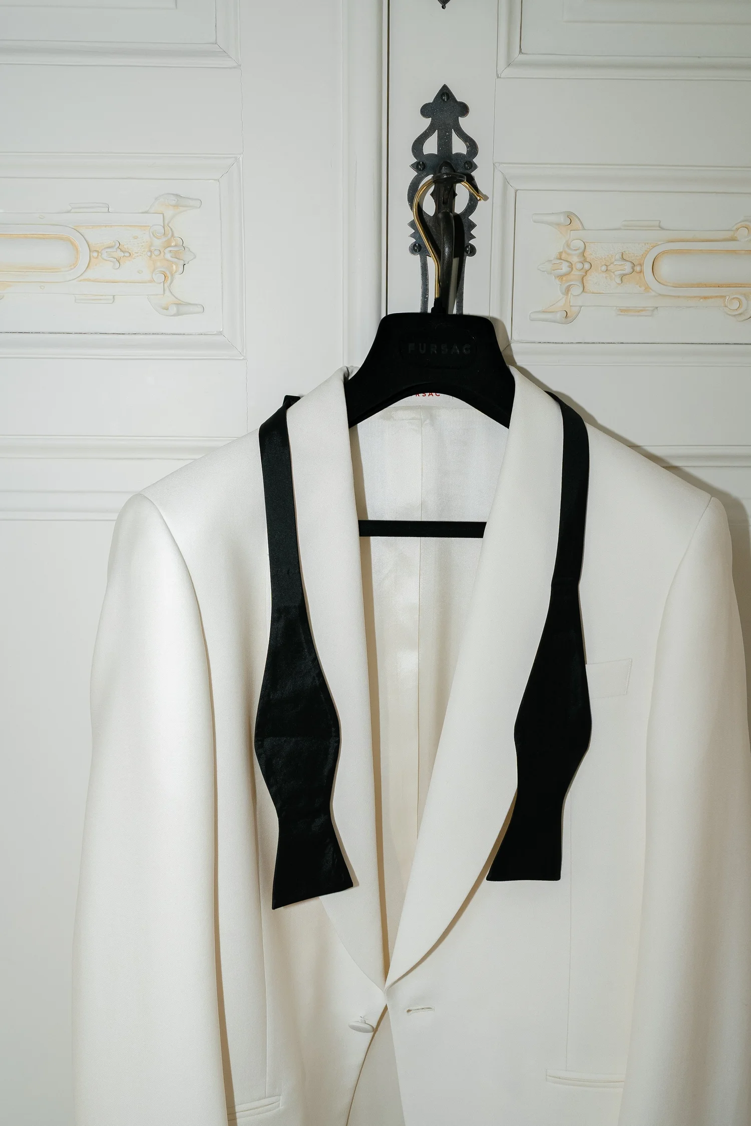 Ivory groom’s dinner jacket and untied black bow tie hanging against ornate white doors