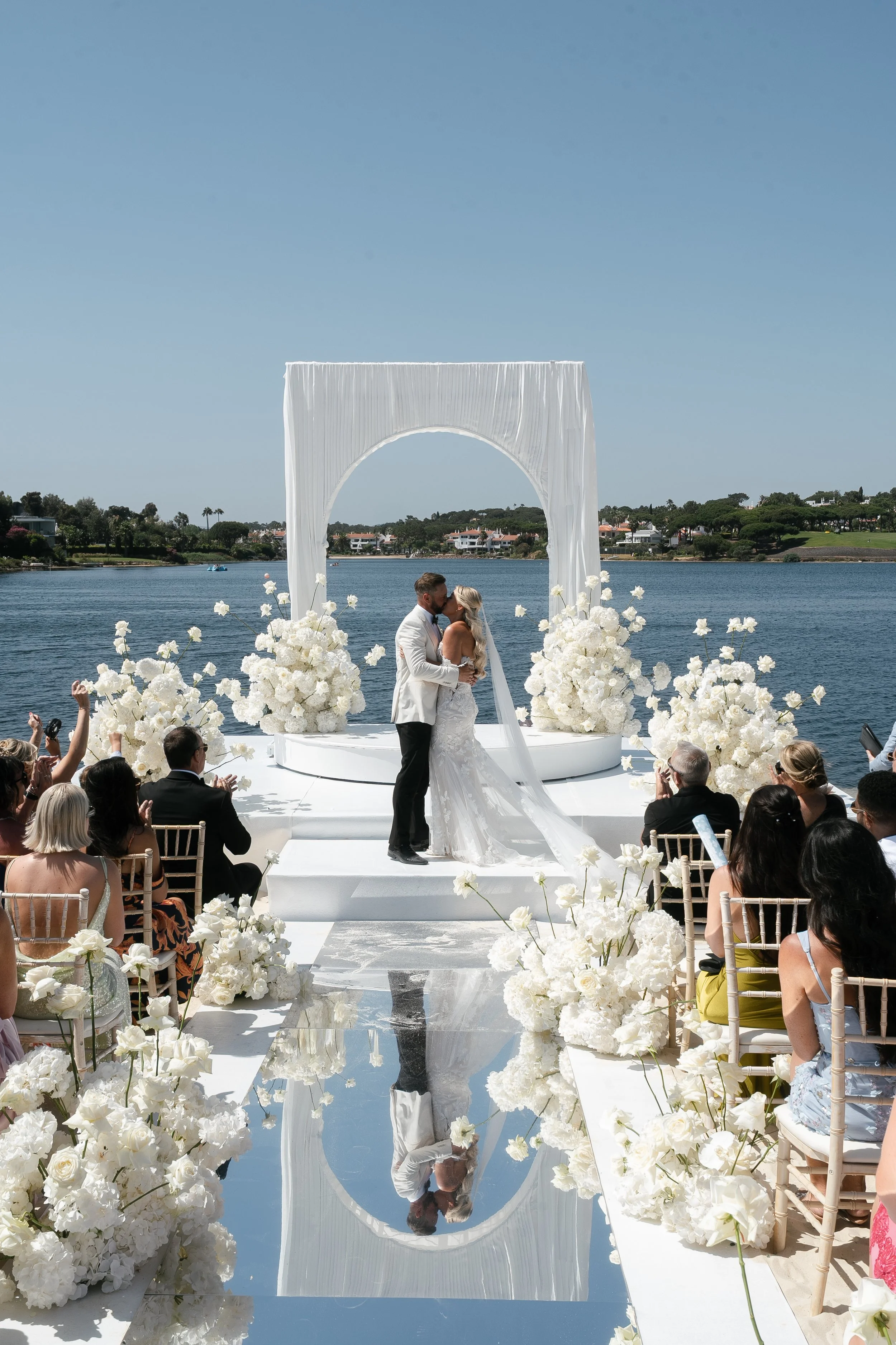 A wedding ceremony by a river with a couple kissing at the altar, surrounded by white flowers and an outdoor audience, under a clear blue sky.
