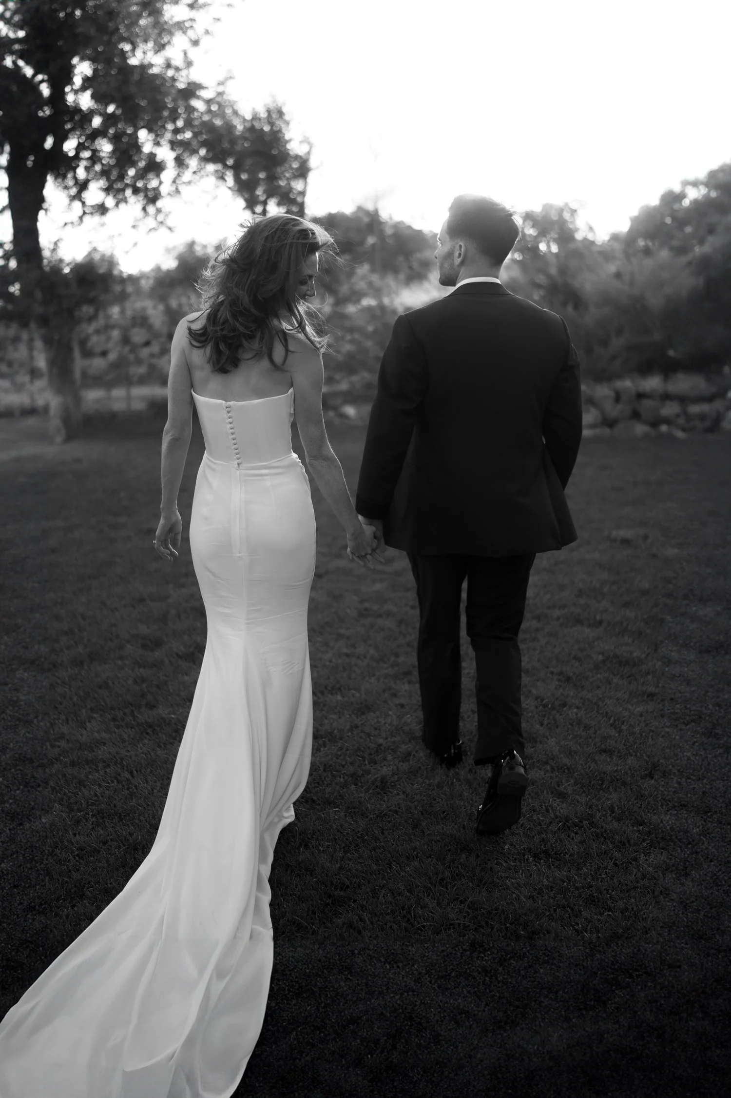 Black-and-white wedding portrait of newlyweds walking hand in hand through a sunlit garden