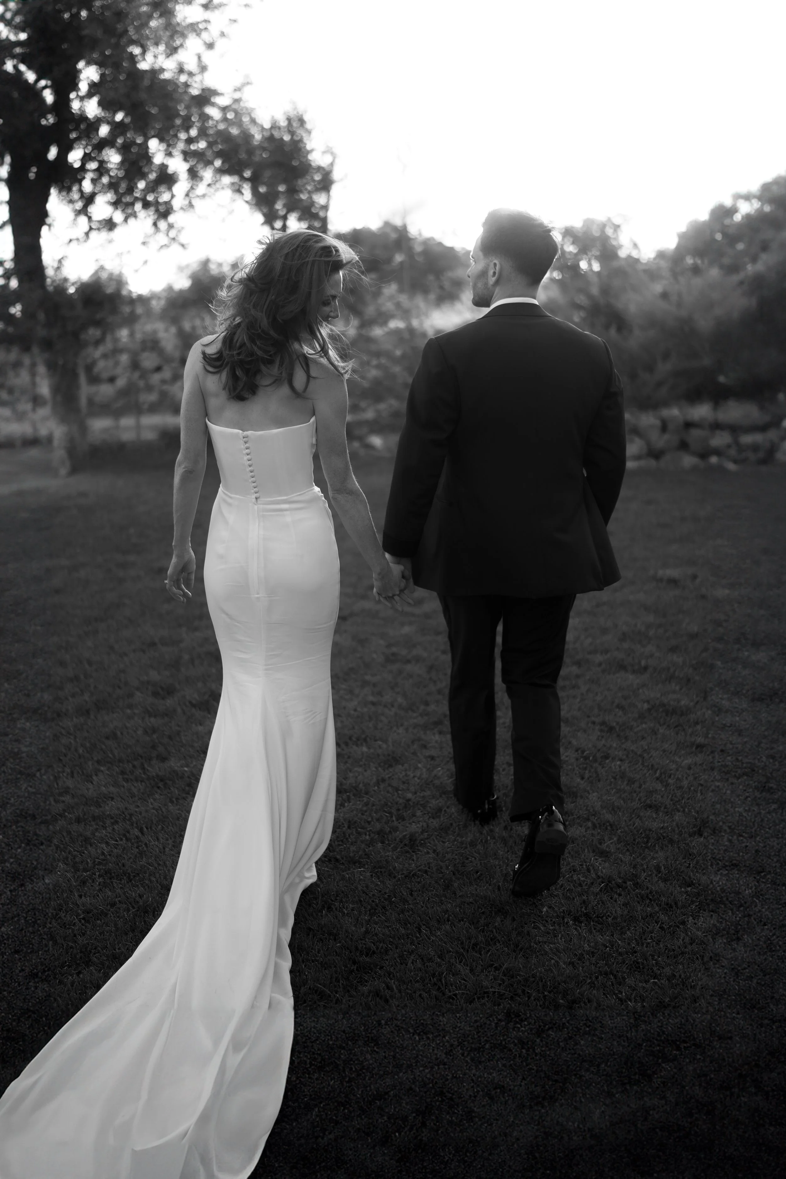 A bride and groom walking hand-in-hand outdoors during sunset, with the bride in a strapless wedding gown with a train and the groom in a black suit.