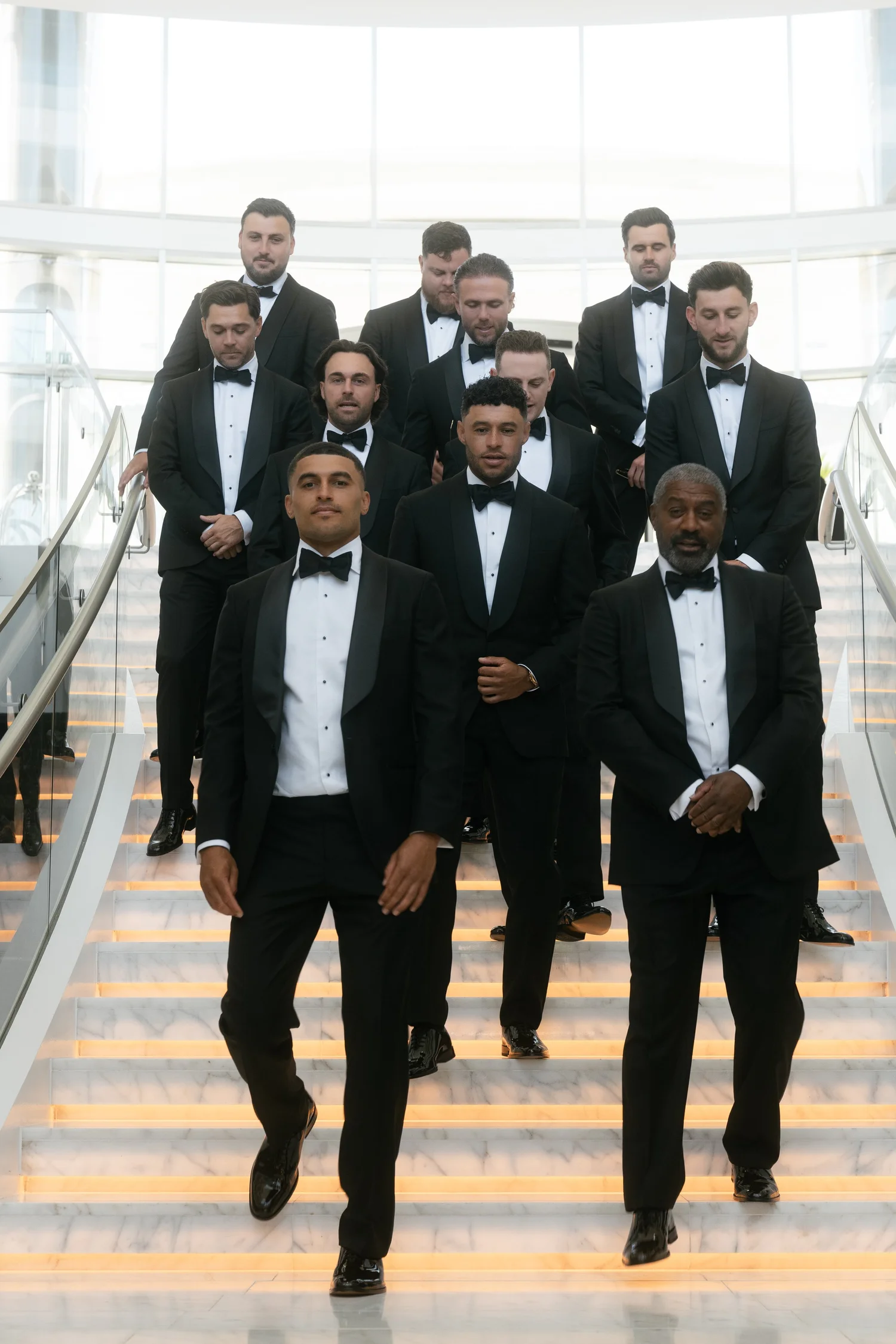 Groom and groomsmen in black tuxedos walking down a bright staircase