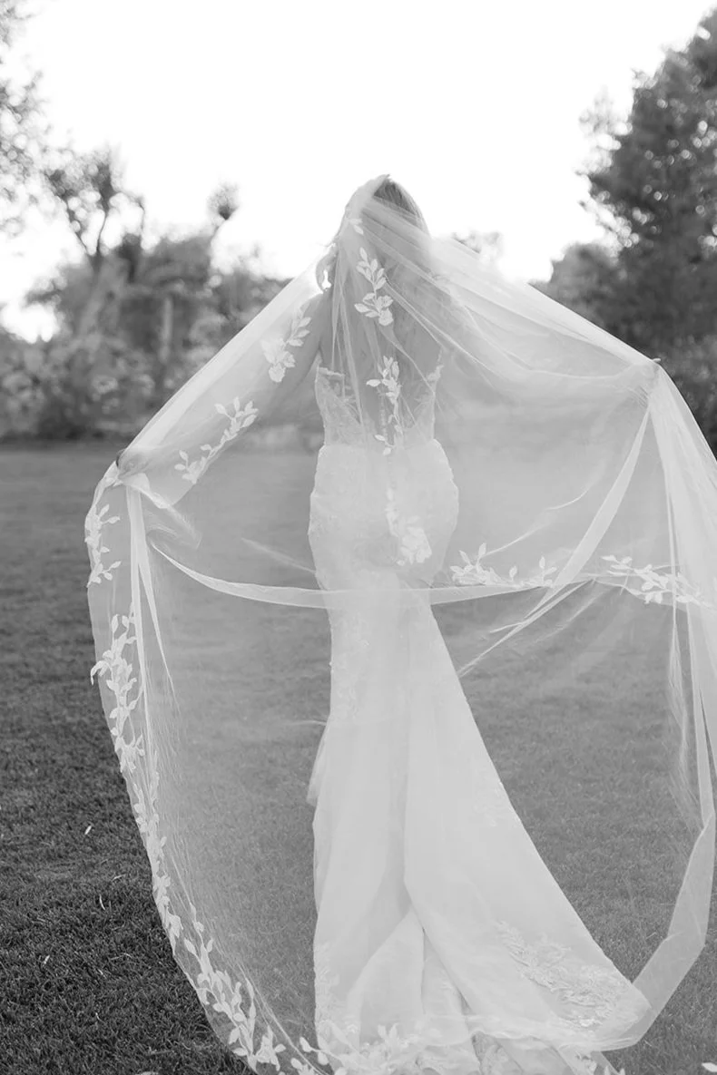 A bride standing outdoors on a grassy area, wearing a wedding dress and a veil with floral embroidery, with trees in the background.