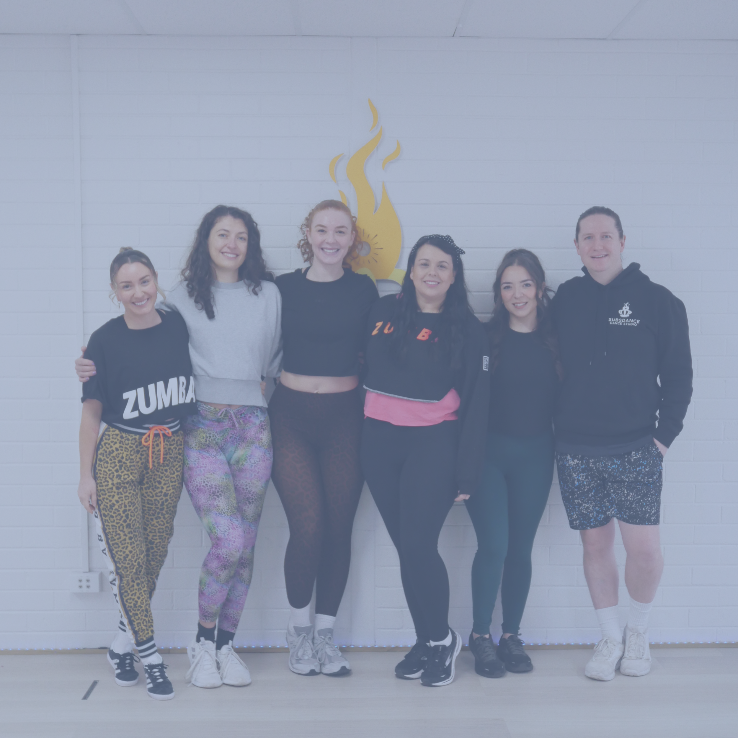 Group of six people standing in front of a white brick wall with a yellow flame logo. They are smiling and dressed in activewear.