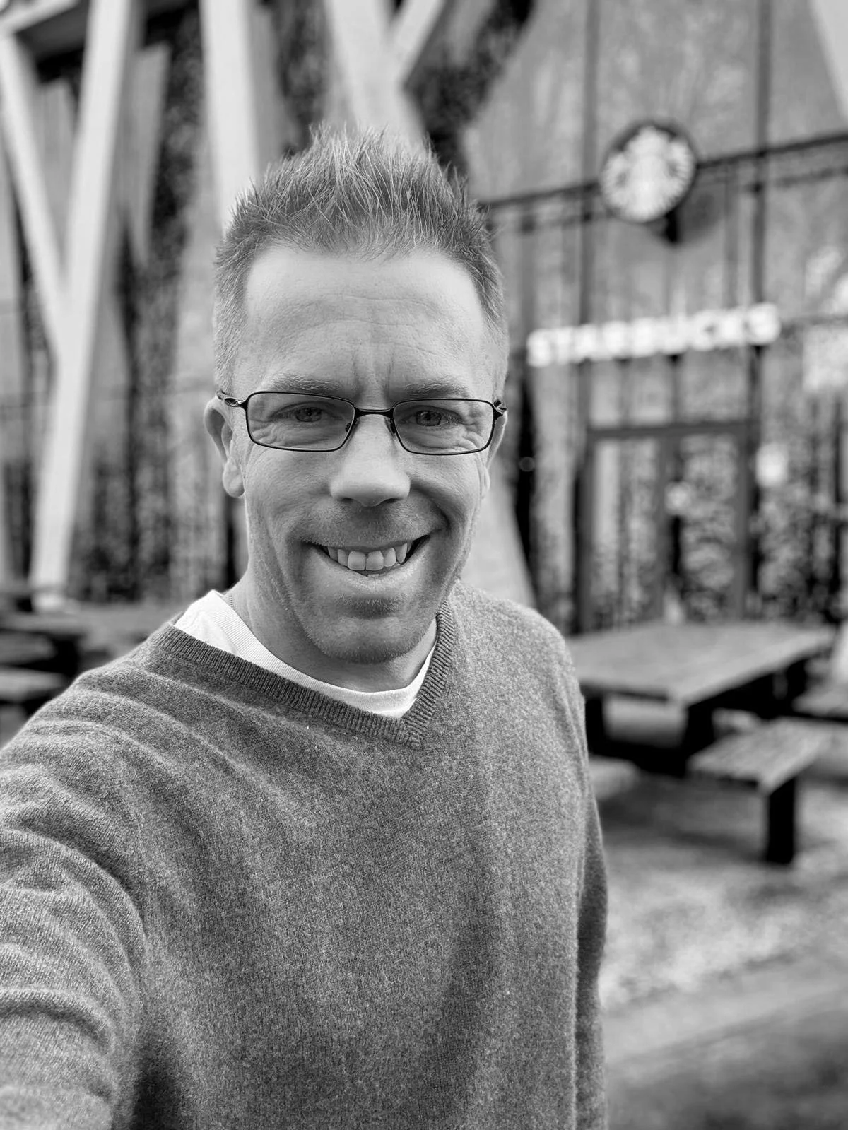 A man smiling for a selfie outdoors, wearing glasses and a sweater, with outdoor furniture and a building in the background.