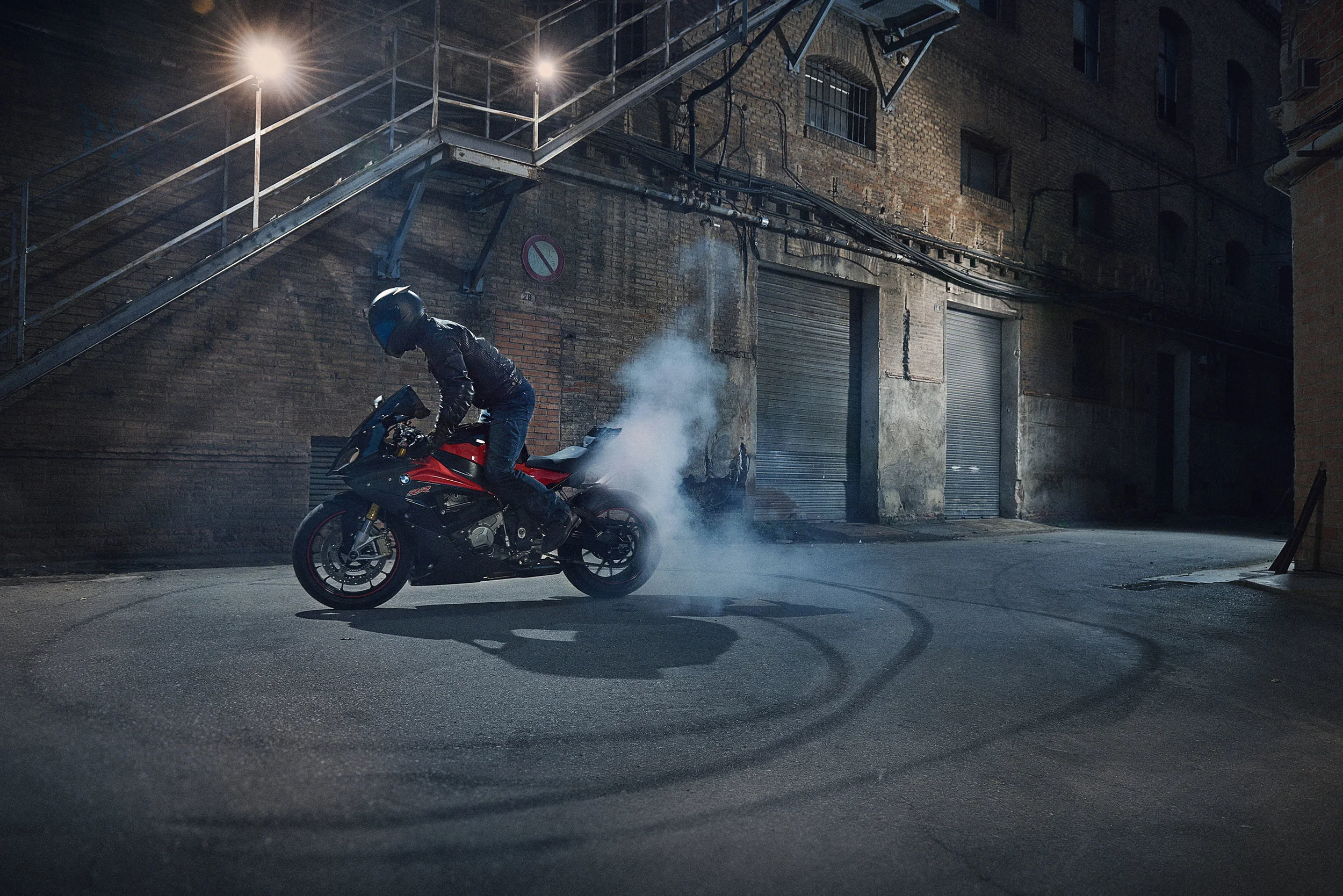 A motorcyclist performing a burnout on a red and black motorcycle in an urban alley at night, with brick buildings and metal staircase in the background.
