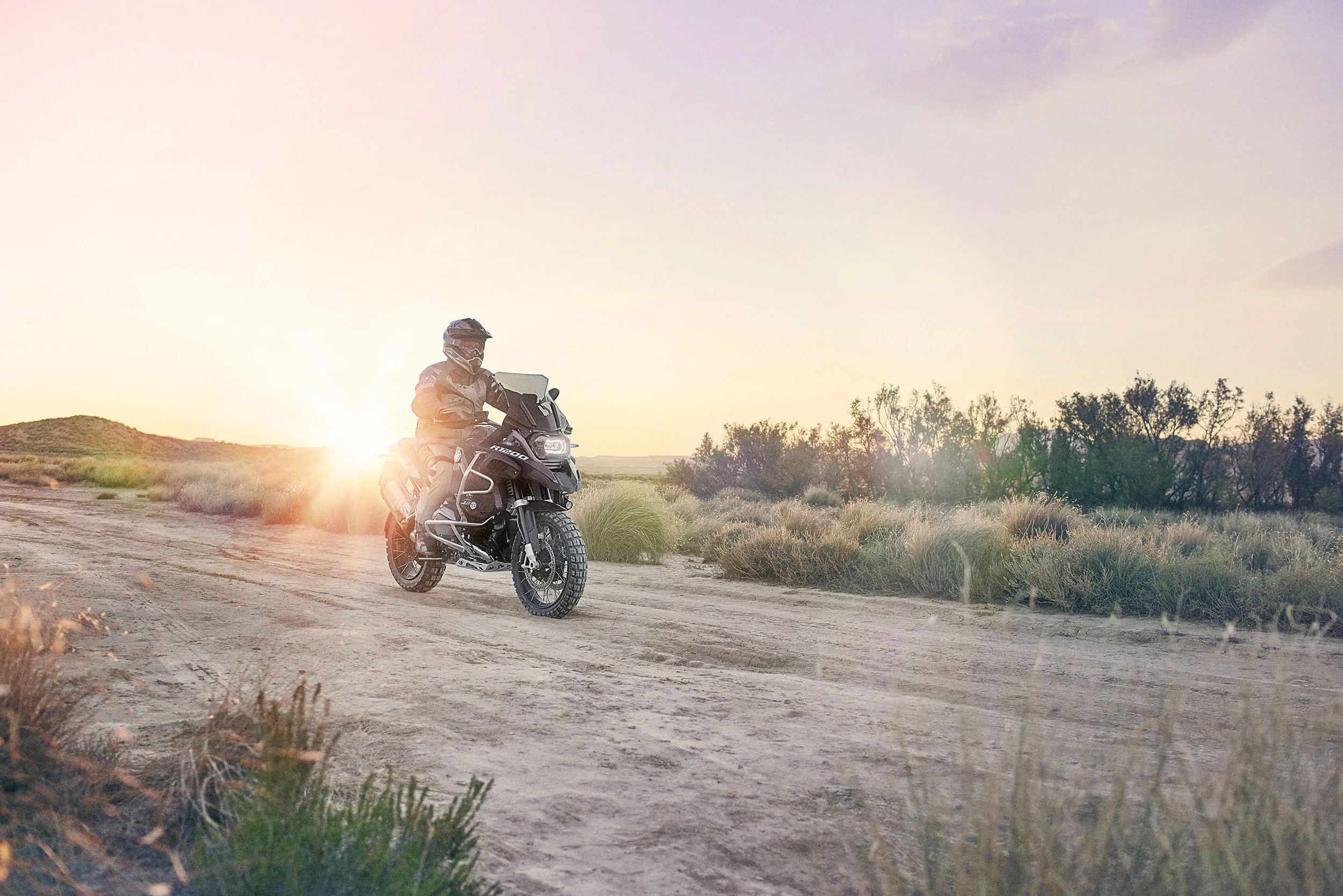 A person riding a motorcycle on a dirt path in a desert landscape at sunset.