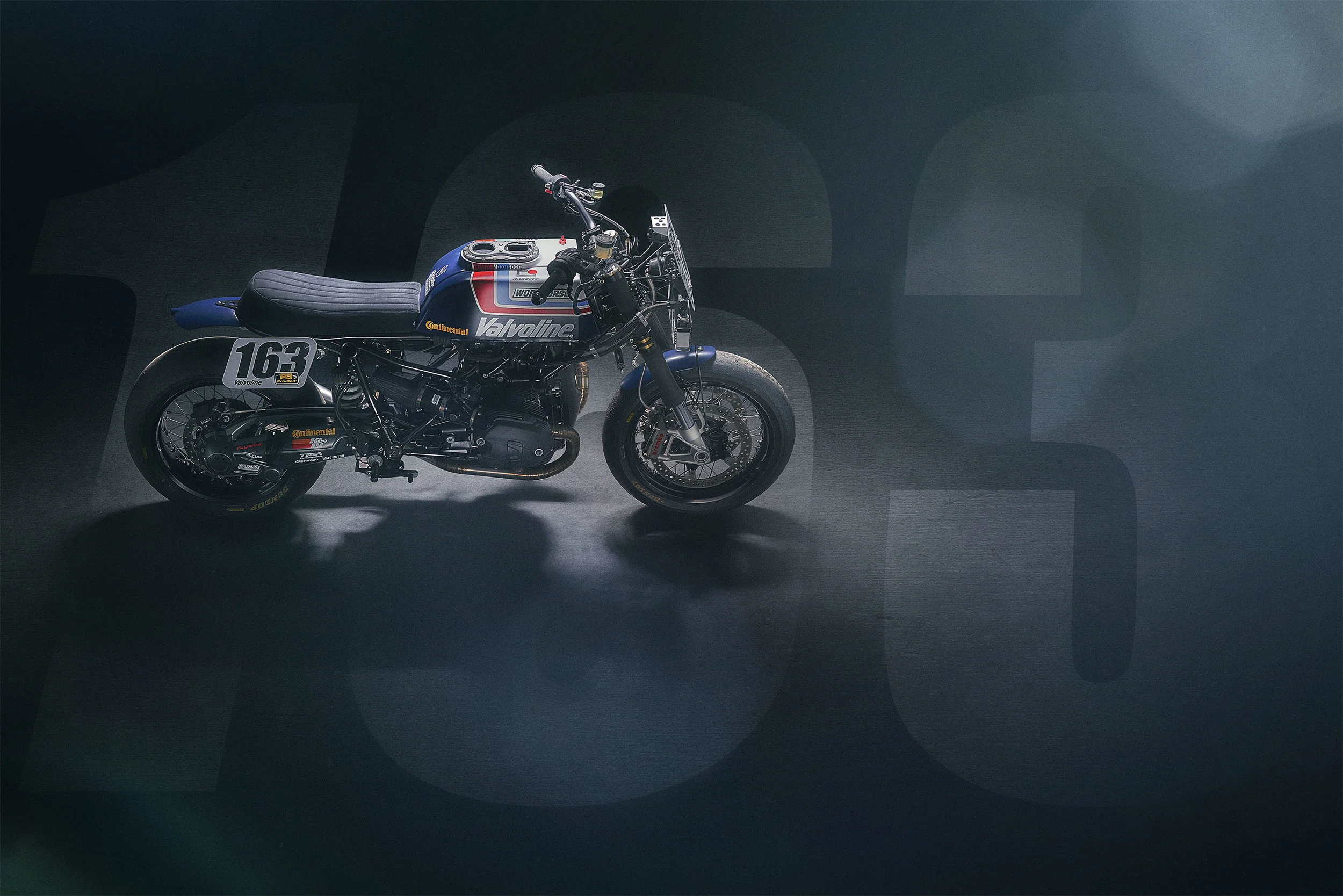 A vintage racing motorcycle with Valvoline branding, number 163, displayed on a dark background with subtle large numbers, and a spotlight highlighting the bike.