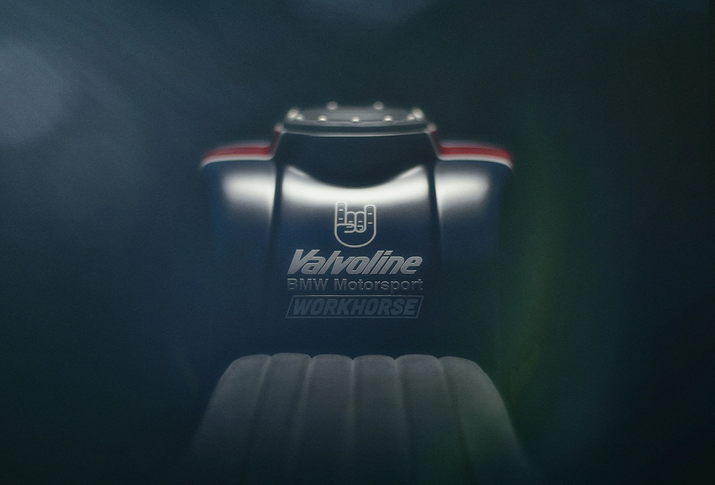 Close-up of a dark racing helmet with red accents, featuring the Valvoline, BMW Motorsport, and Workhorse logos, displayed against a dark background with a reflective surface.