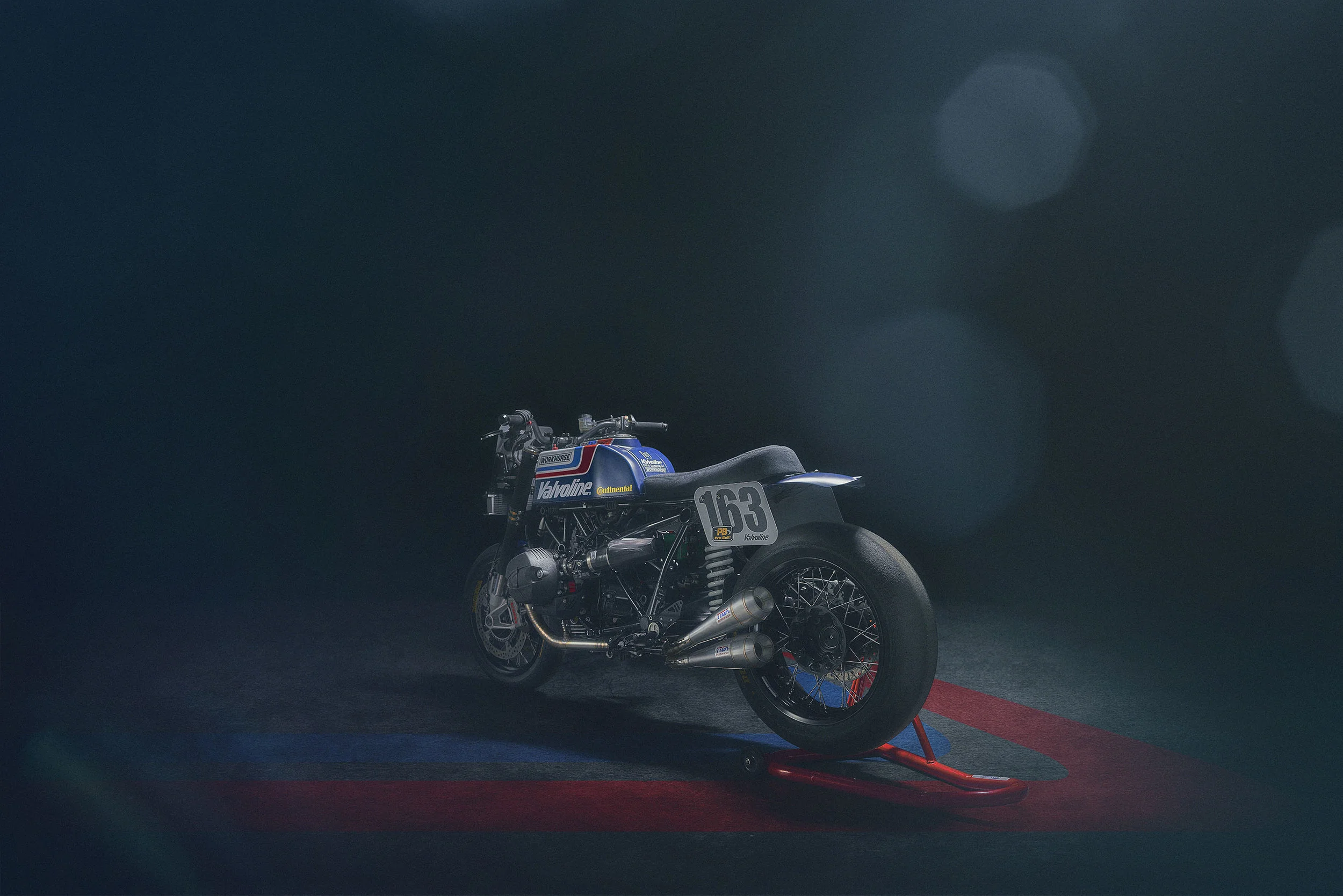 A vintage race motorcycle, number 163, on a dark background with subtle light effects, resting on a red stand over a blue and red striped mat.
