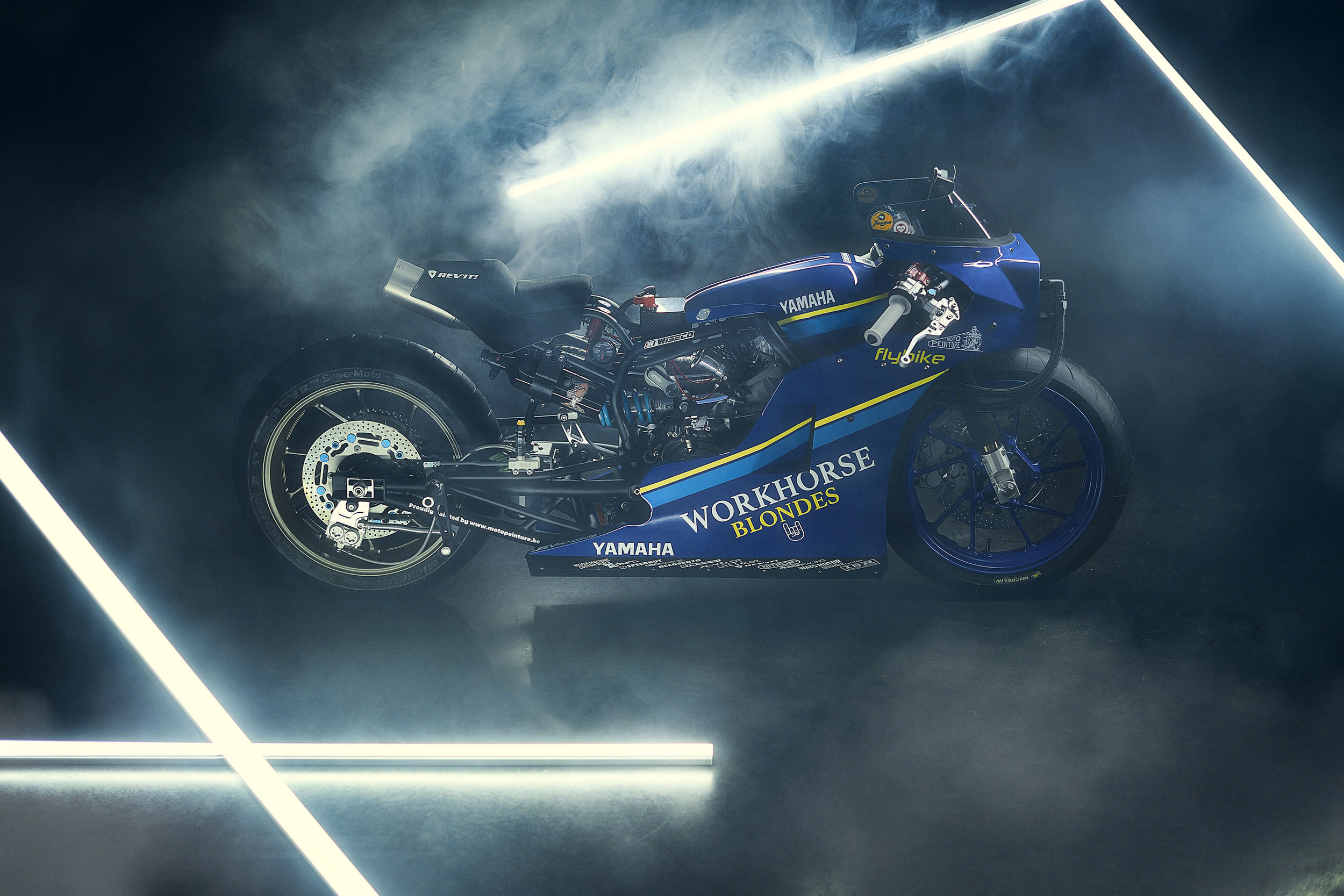 A vintage Yamaha motorcycle with racing decals and sponsorships is displayed in a neon-lit foggy setting.