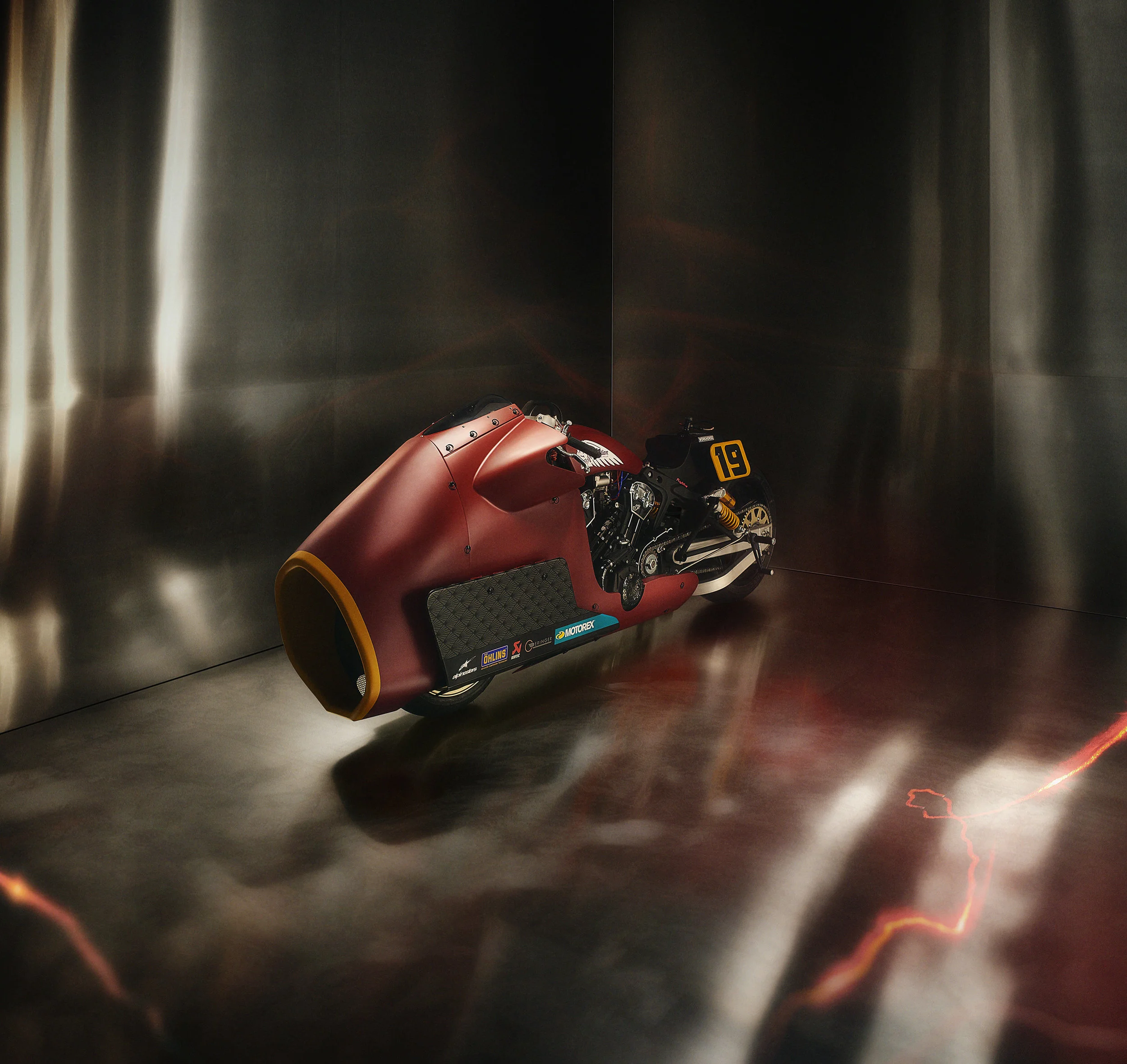 A sleek, modern racing motorcycle with a matte red aerodynamic fairing displayed in a reflective metallic room.