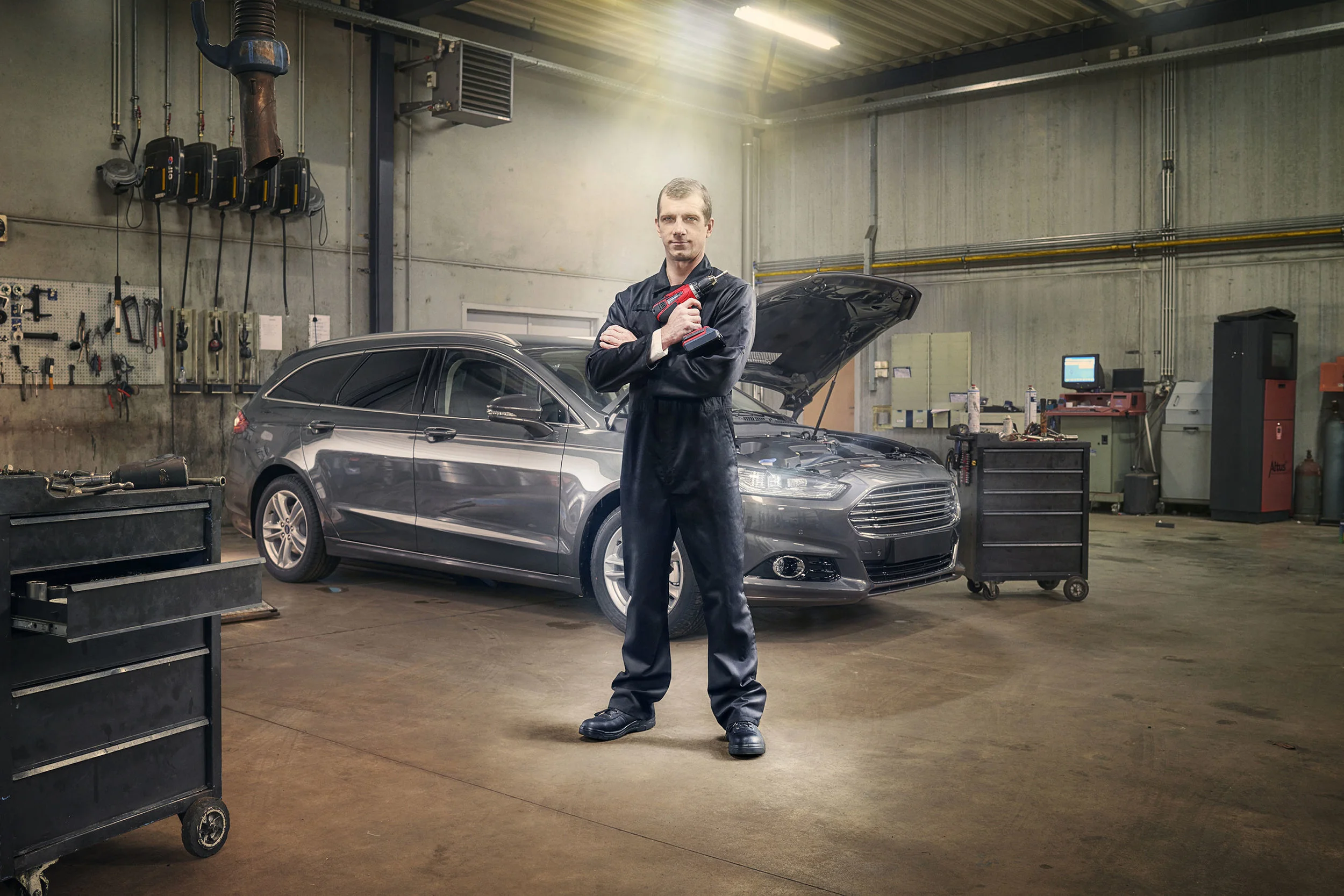 A mechanic standing with arms crossed holding a tool in an auto repair shop next to a silver car with the hood open.