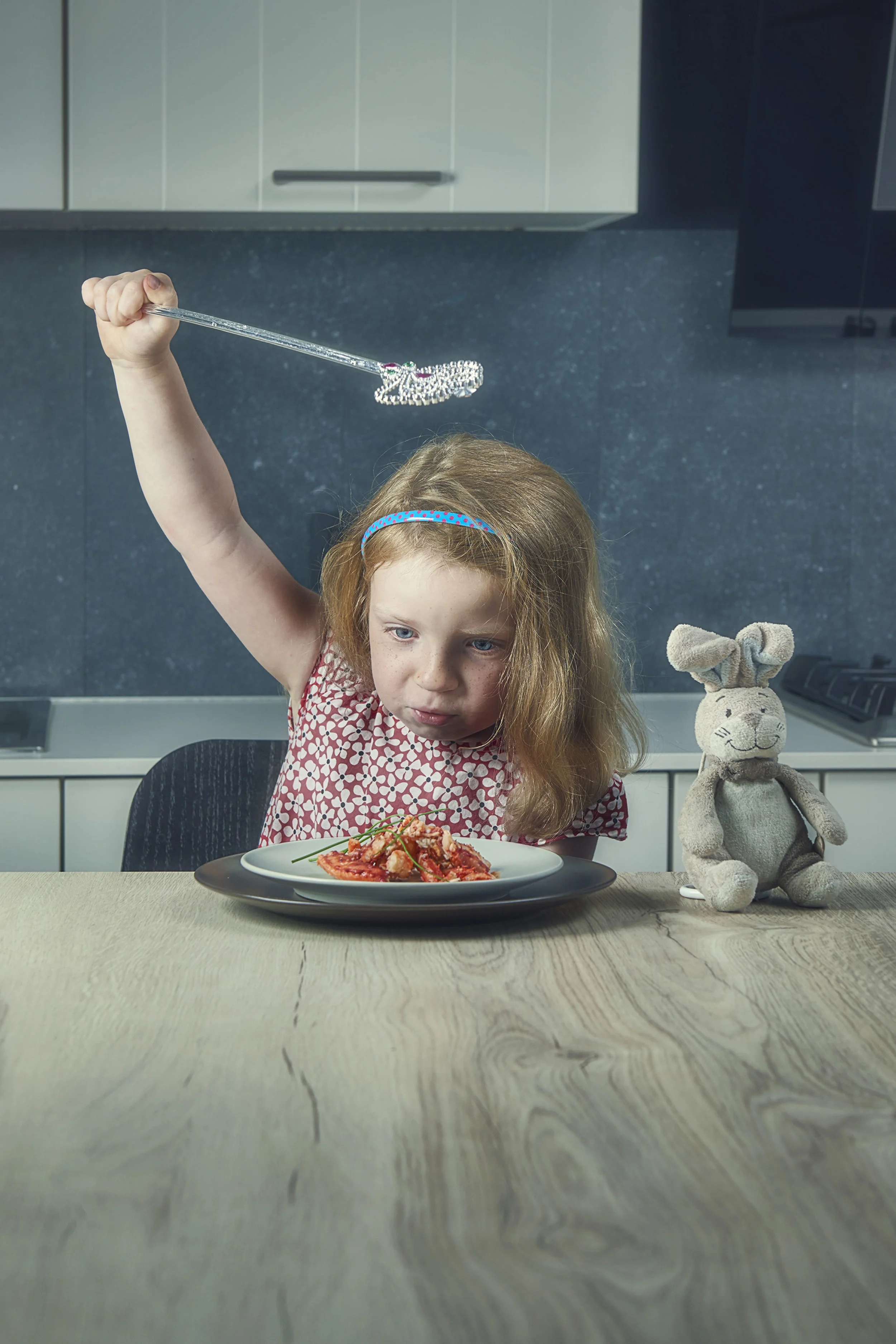 A young girl with red hair and a headband is sitting at a kitchen table, raising a long silver utensil and looking at a plate of spaghetti with tomato sauce. On the table to her right is a stuffed rabbit toy.
