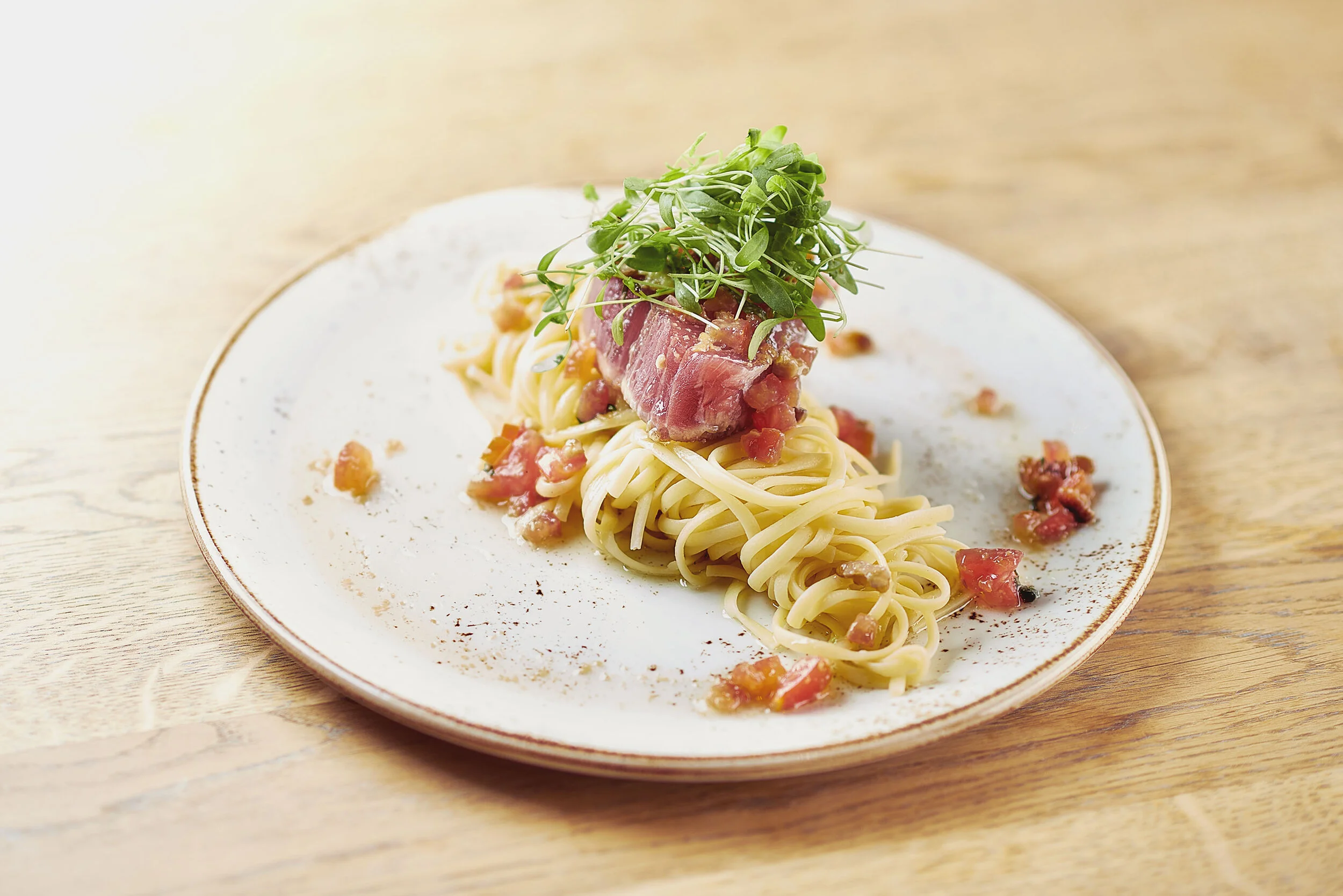 Plate of pasta with raw tuna, tomato, and microgreens on top, served on a white plate on a wooden table.