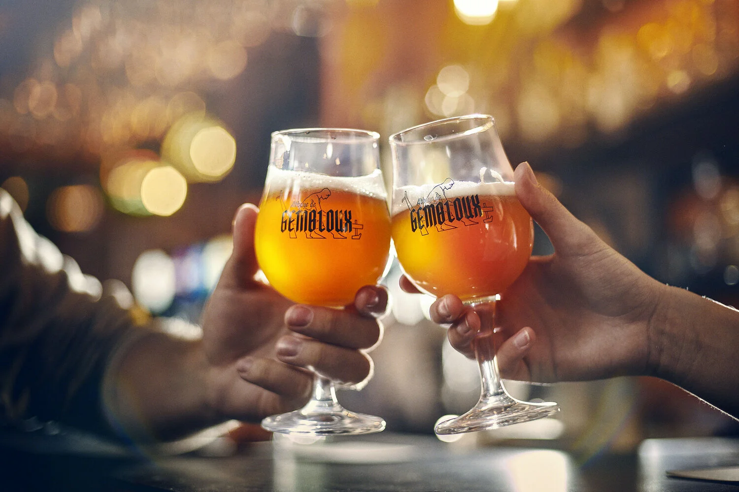 Two people clinking glasses of beer in a bar with warm bokeh lights in the background.