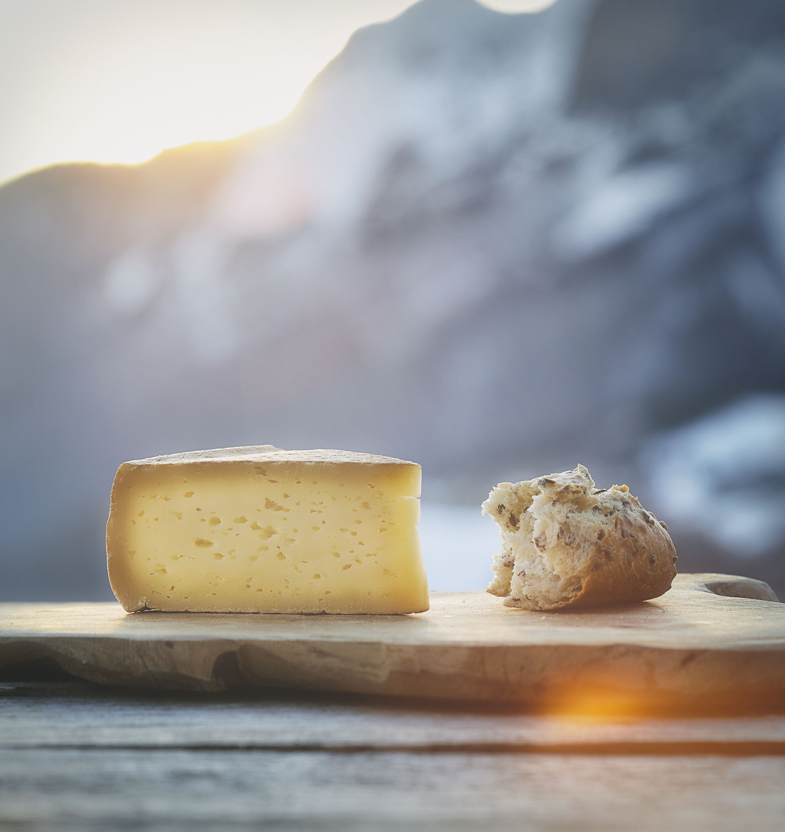 A wedge of yellow cheese and a piece of bread on a wooden cutting board with a snowy mountain landscape in the background.