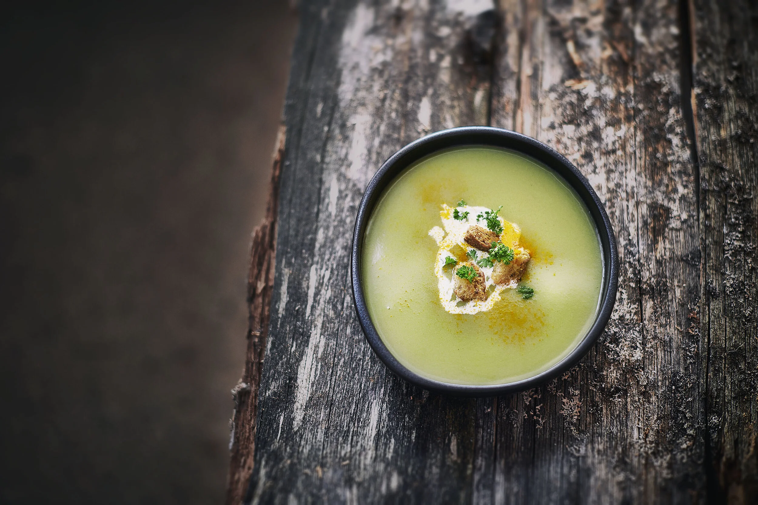 A bowl of green soup garnished with herbs, croutons, and a dollop of cream, placed on a weathered wooden surface.