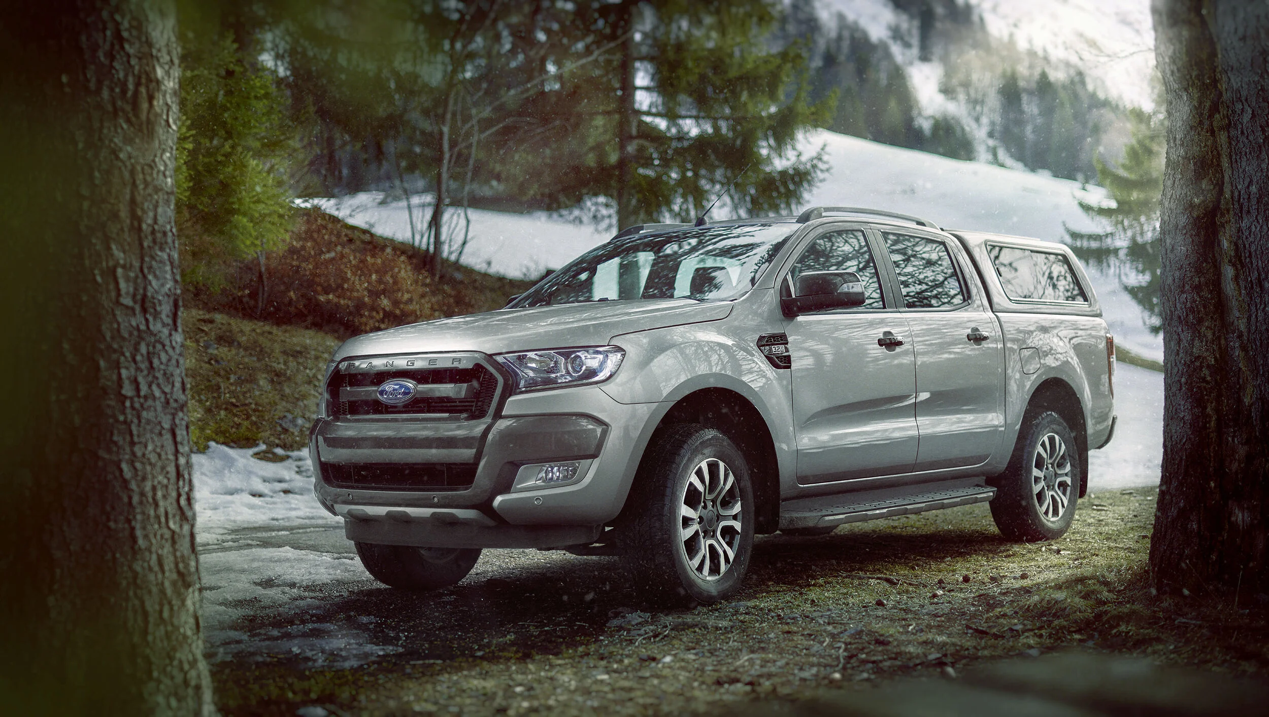 A silver Ford Ranger pickup truck parked in a forested area with trees, snow, and a dirt ground.