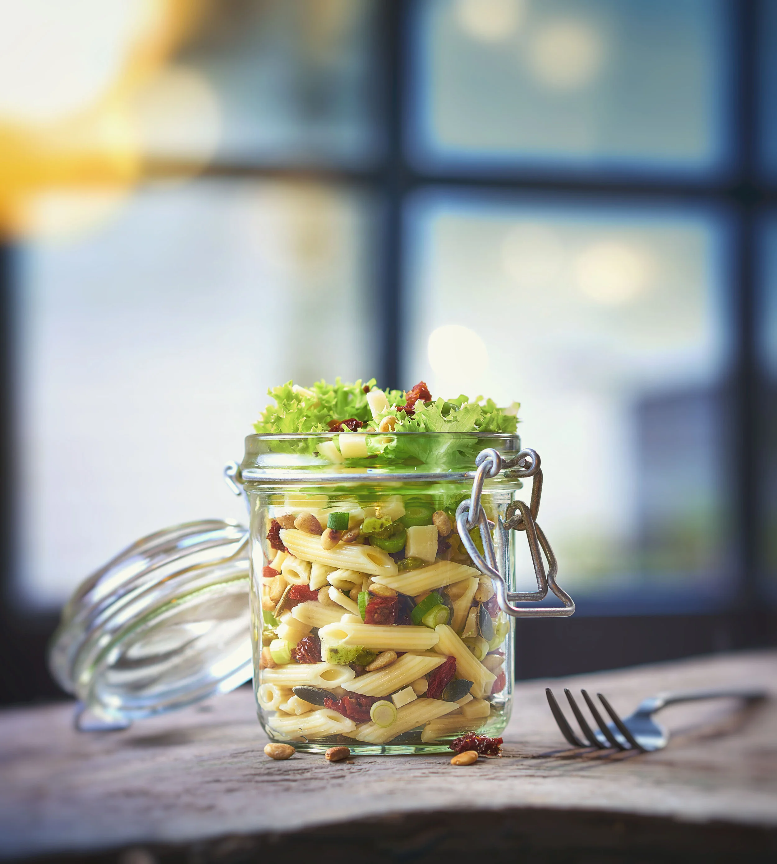 A glass jar with a metal clasp lid containing pasta salad with penne, green onions, sun-dried tomatoes, and mixed greens, placed on a wooden surface with a fork and some scattered ingredients, against a blurred window background.
