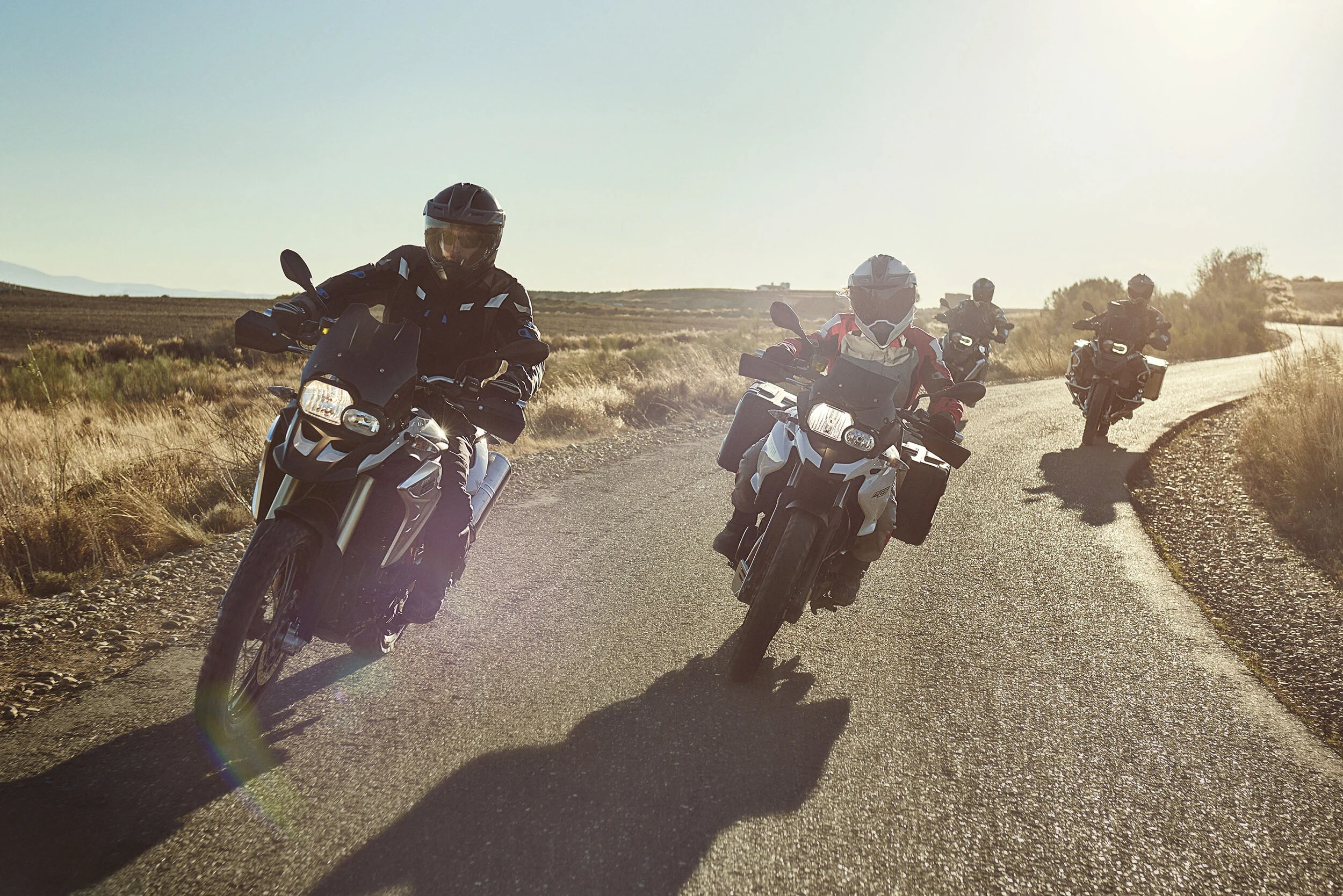 Four motorcyclists riding on a winding rural road during sunset, with open fields and bushes on either side.