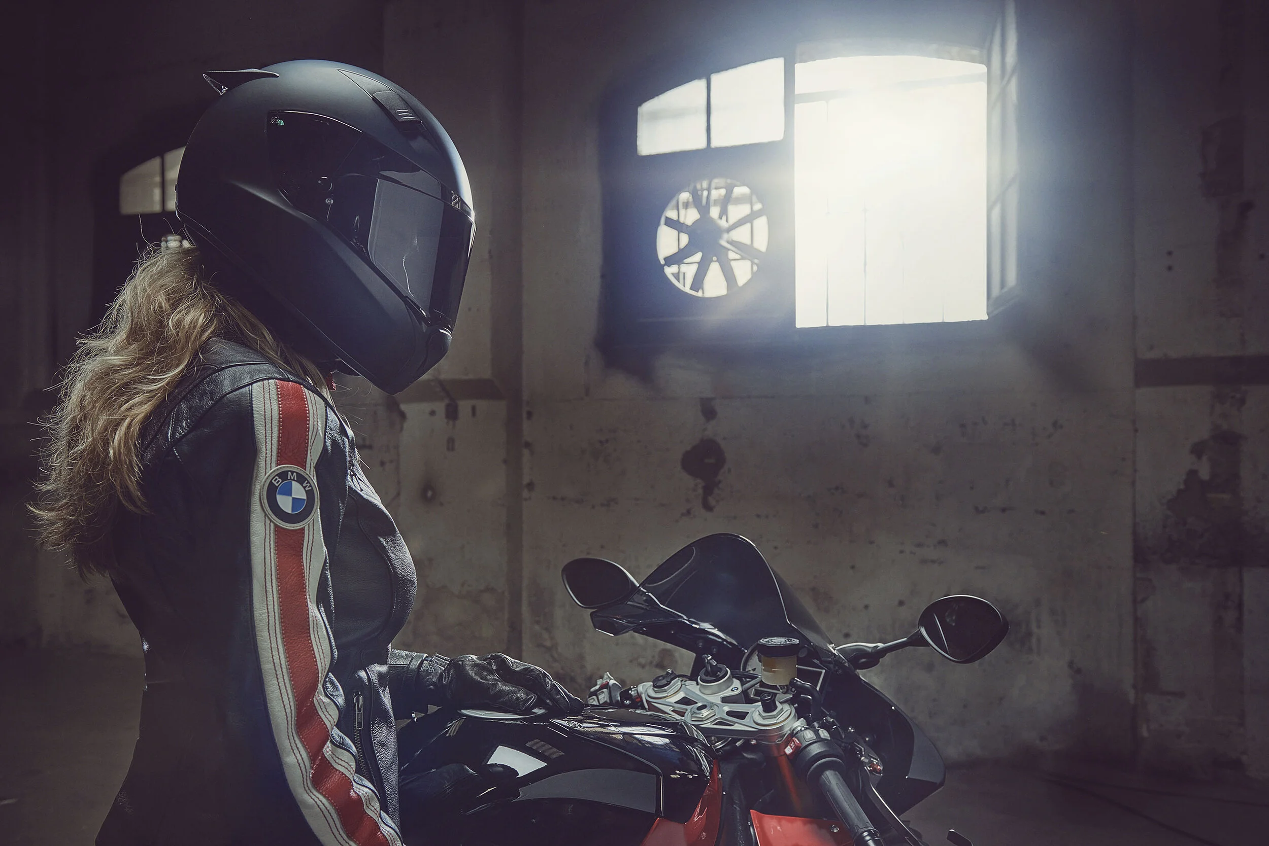A person with long curly hair wearing a BMW motorcycle jacket and a full-face black helmet, standing next to a black motorcycle inside a dimly lit industrial-style room.
