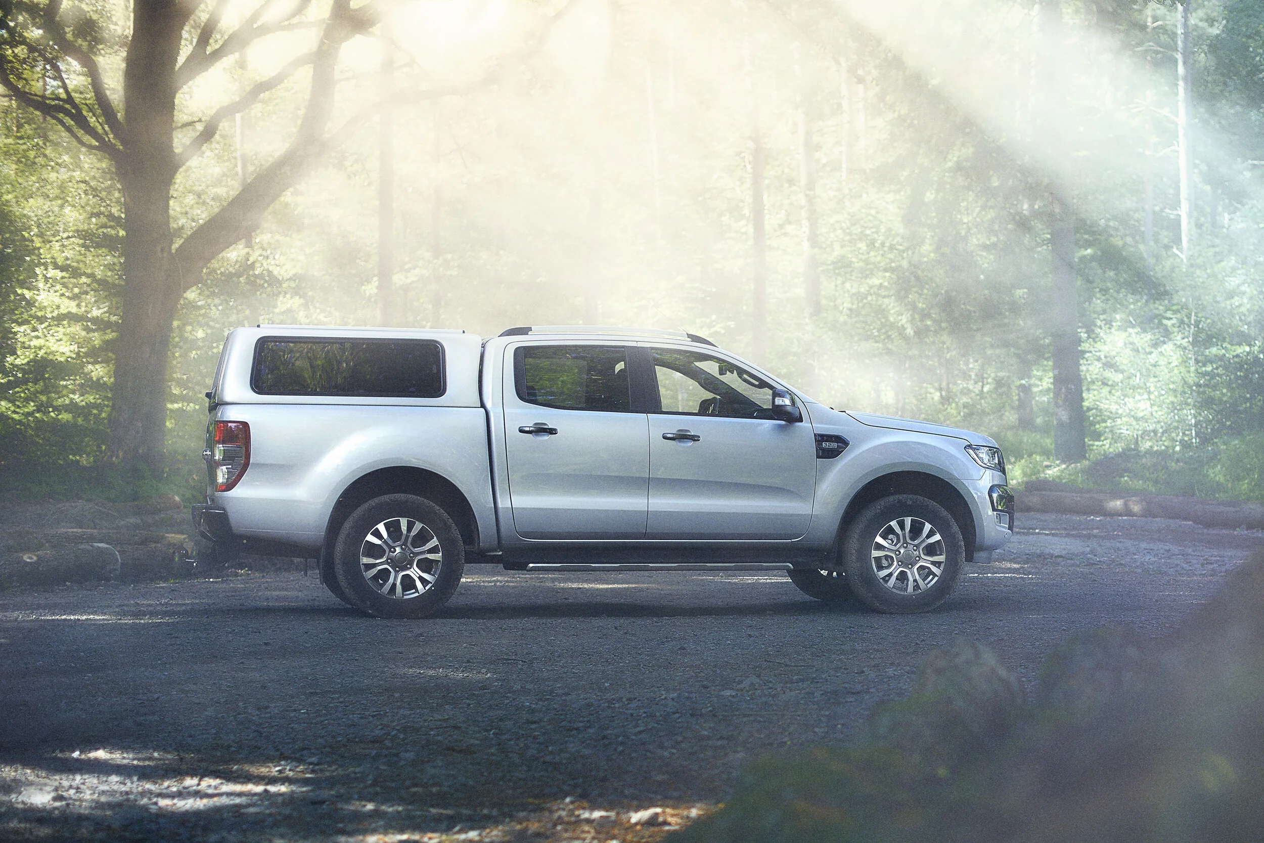 A silver pickup truck parked on a dirt road in a forest with sunlight filtering through the trees.