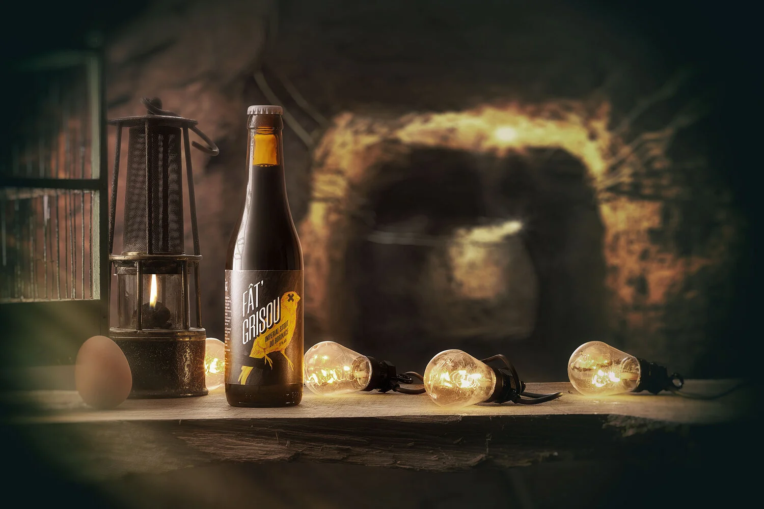 A bottle of beer, an egg, a vintage lantern, and string lights on a wooden surface in a rustic setting.