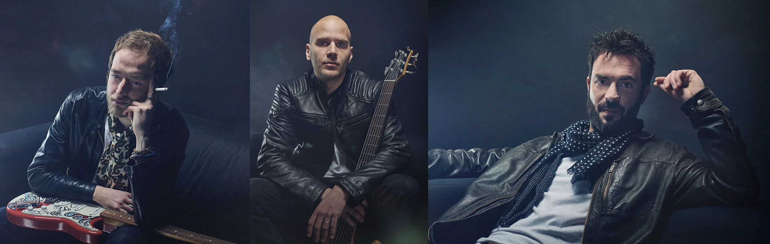 Three men in leather jackets sitting on sofas against a dark background, each with a guitar.