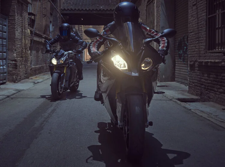 Two motorcyclists wearing helmets riding bikes down a narrow urban alleyway surrounded by brick buildings.
