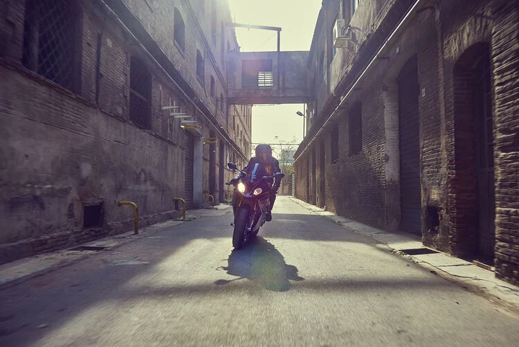 A person riding a motorcycle down a narrow urban alleyway with brick buildings on either side and a bridge overhead, sunlight shining from behind.