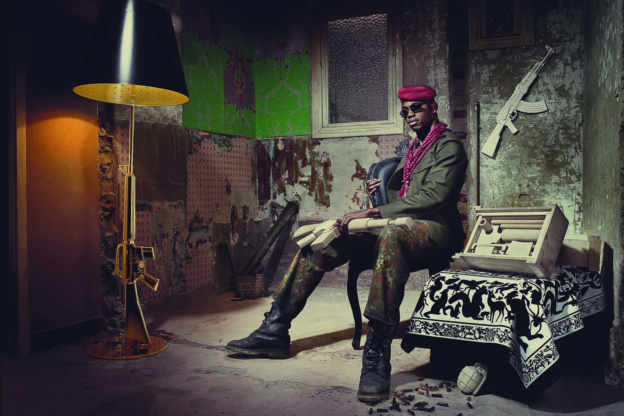 A young man sitting in a run-down room with graffiti-covered walls, wearing sunglasses and a maroon beret, surrounded by fake weapons and military paraphernalia, with a black-and-white patterned tablecloth and scattered bullets on the floor.