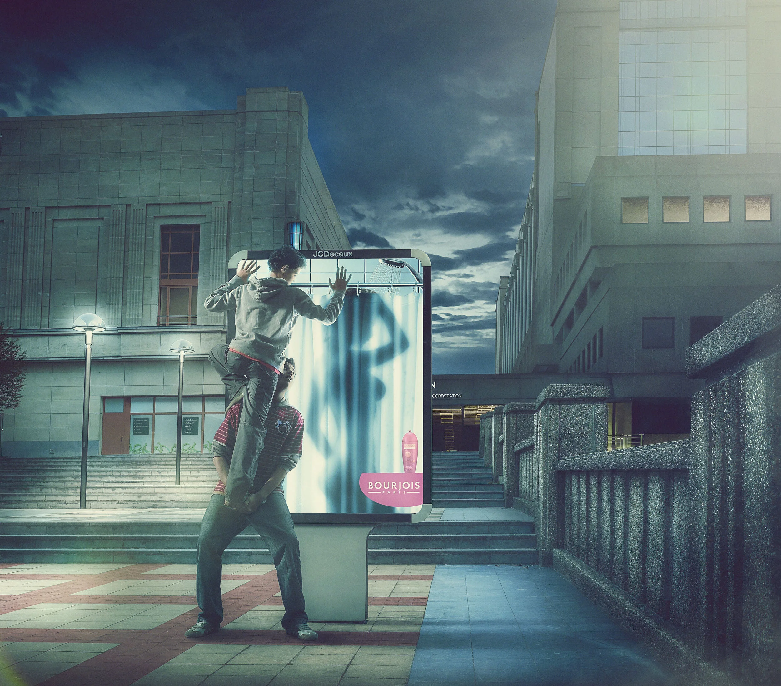 Two young men, one standing on the other's shoulders, appear to be hiding behind or in front of a large advertisement display in an urban setting at dusk, with modern buildings and street lamps in the background.