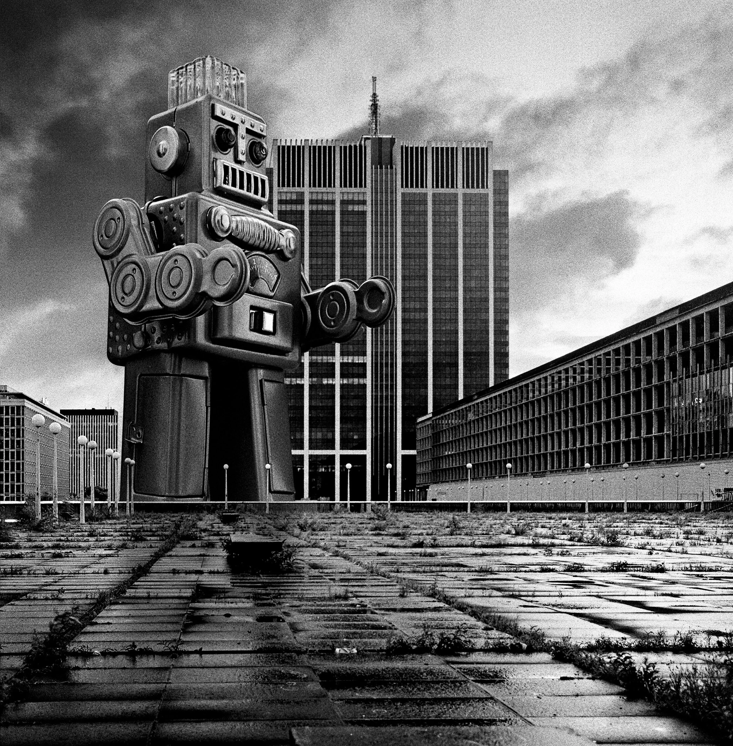 A black and white image of a giant robot sculpture in front of a tall, modern office building in an urban setting, with a cloudy sky overhead.