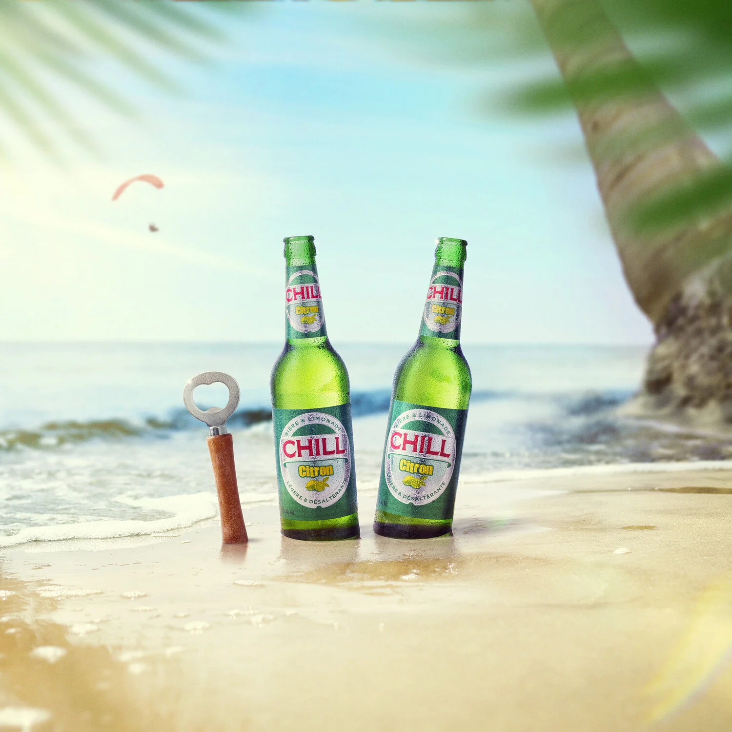 Two green bottles labeled 'Chill Citron' standing on the sandy beach near the shoreline with a small bottle opener placed next to them. In the background, the ocean waves and a parachute in the sky are visible, with a palm tree on the right side fram