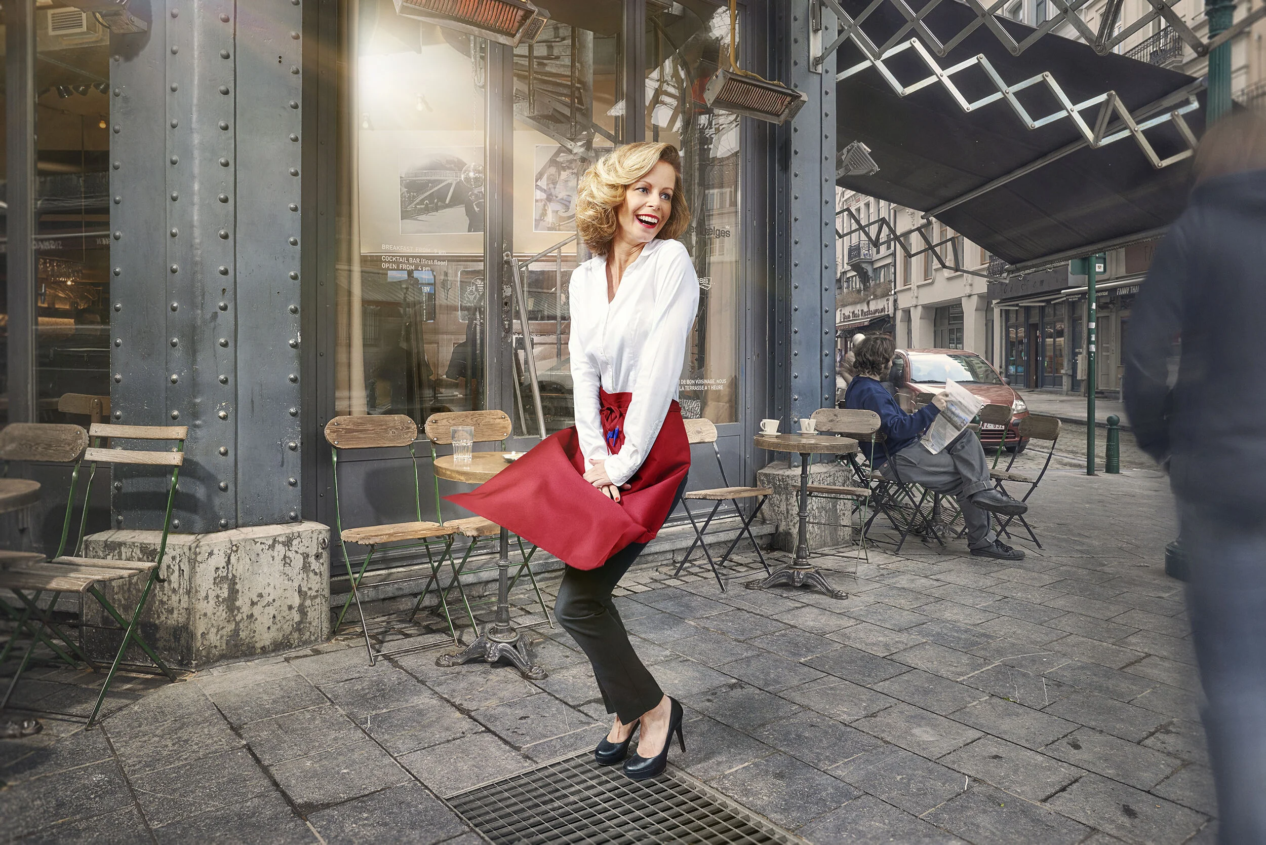 A woman in a white blouse and black pants with a red apron is standing on a city sidewalk outside a cafe, smiling and posing for the camera. She is wearing high heels, and there are empty chairs and tables behind her. A man is sitting on one of the t