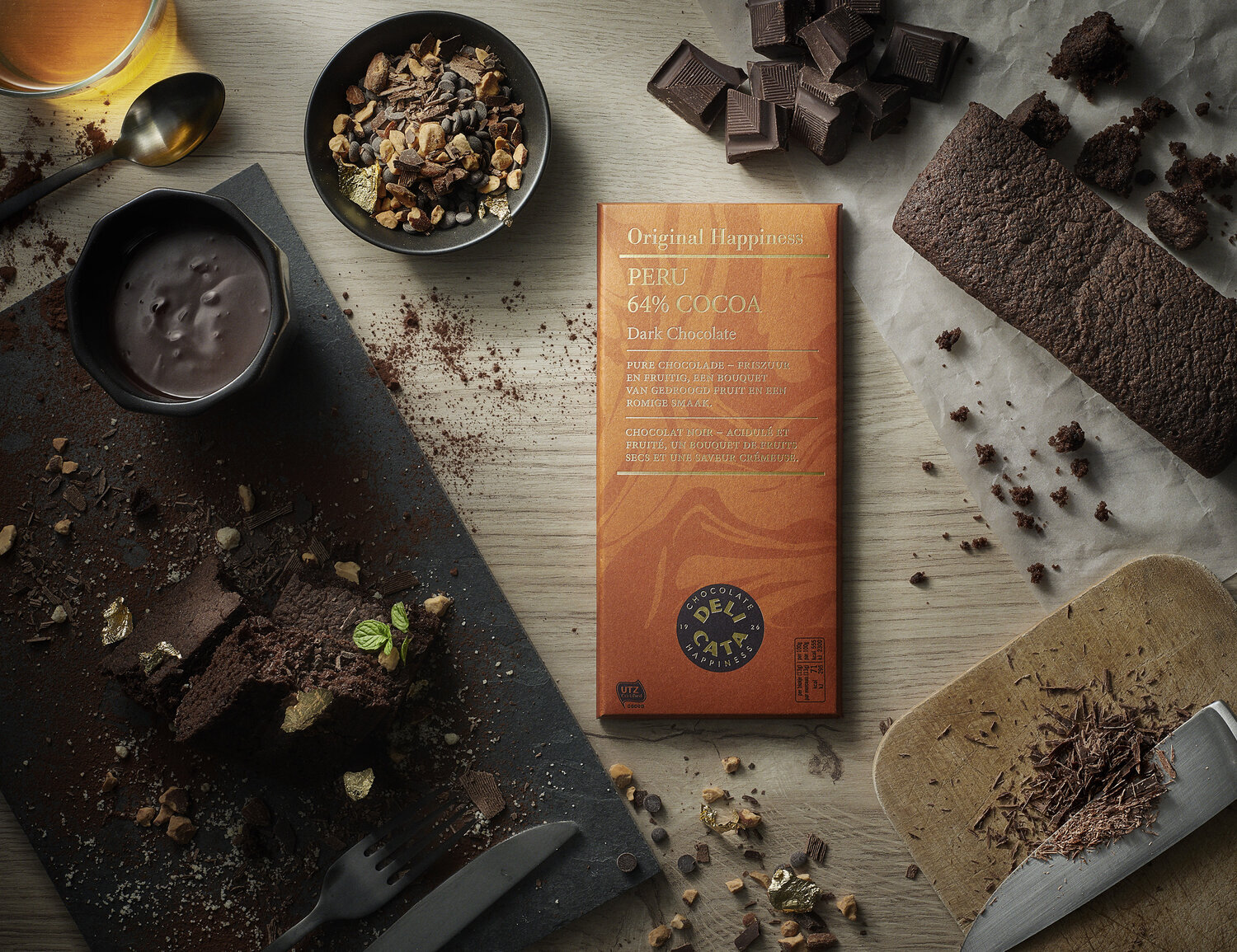 A baking scene with a bar of Peru 64% cocoa dark chocolate, a chocolate cake with gold accents and mint leaves, chopped chocolate pieces, a bowl of mixed nuts, a cup of hot chocolate, and chocolate shavings on a wooden table.
