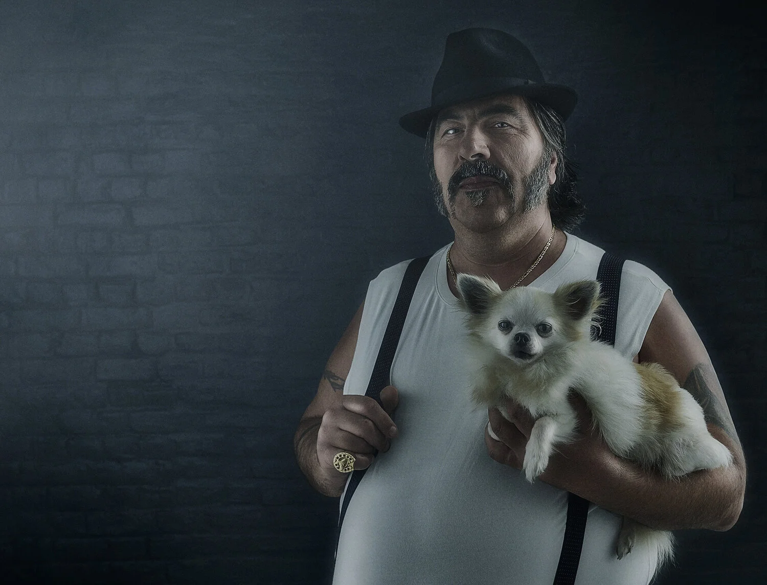 A man with a mustache and beard wearing a black fedora, white sleeveless shirt, and suspenders holds a small fluffy dog with light fur in front of a dark textured background.
