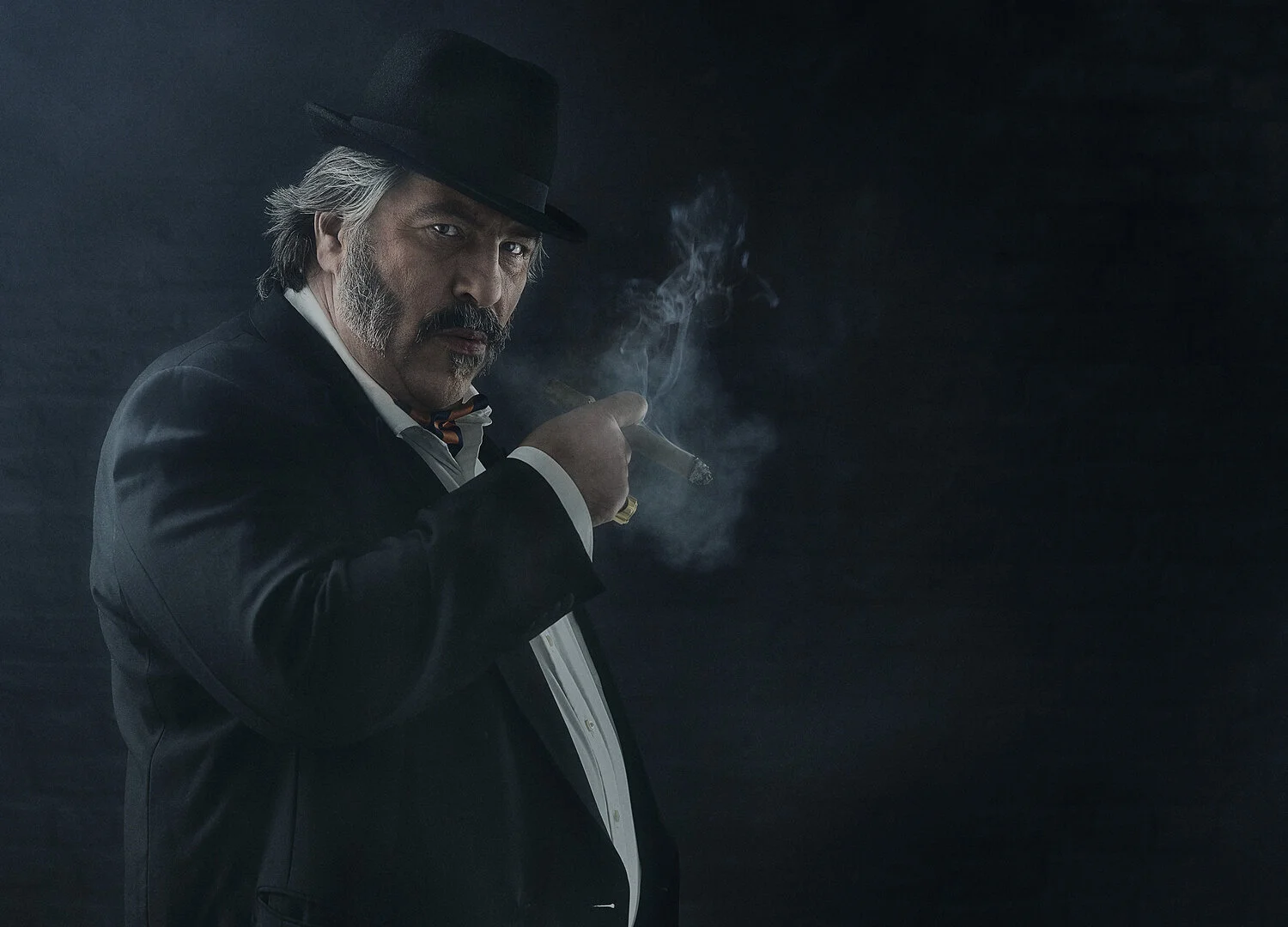 A man with gray hair and a beard, dressed in a tuxedo and black hat, holding a cigar and looking intensely at the camera. The background is dark.