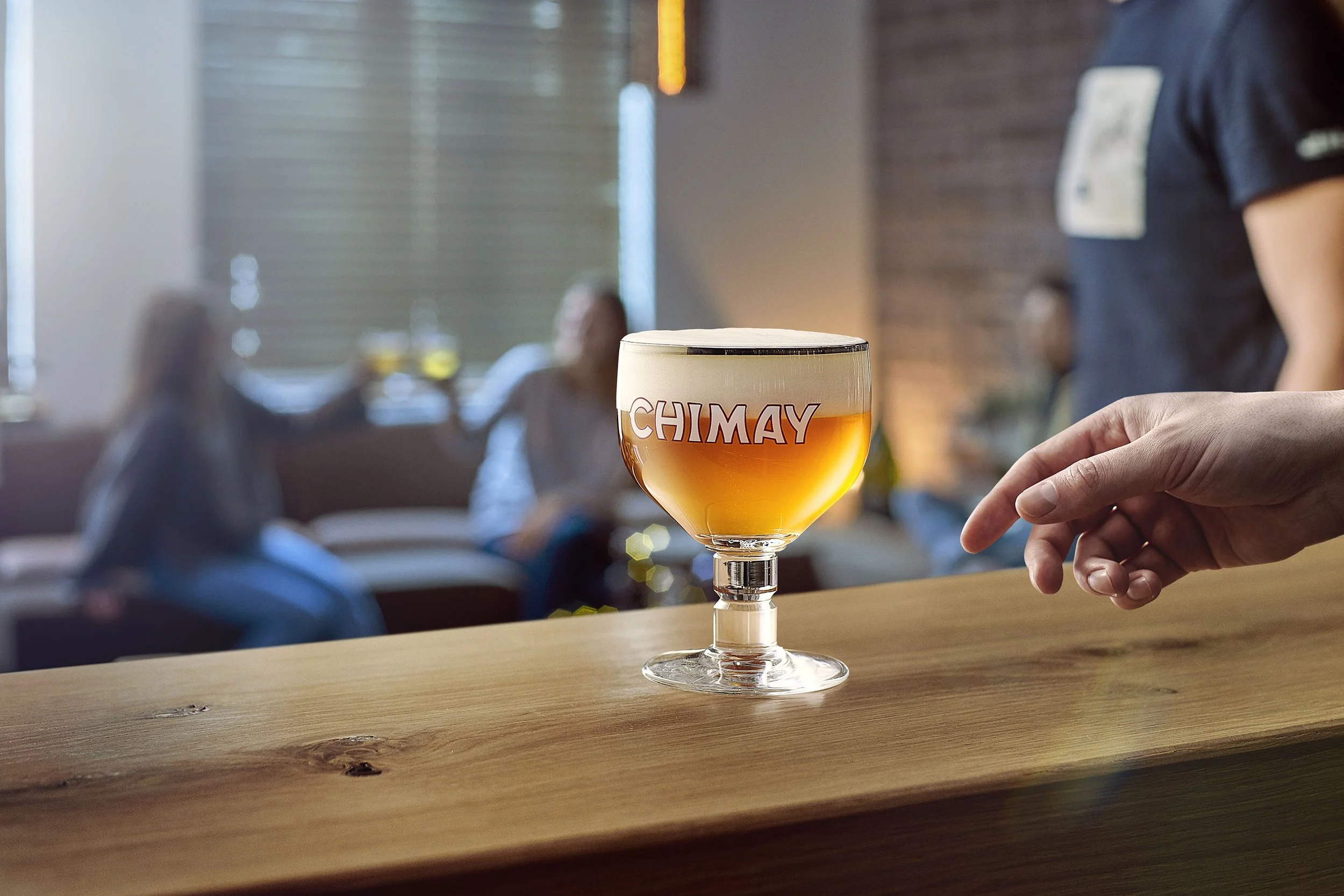 A glass of beer with the label 'Chimay' on a wooden bar, with a person's hand reaching towards it in a lively bar or pub setting.