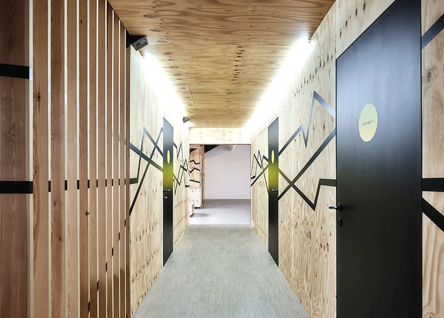 Wood-paneled hallway with black doors featuring green and yellow geometric designs, illuminated by bright ceiling lights.