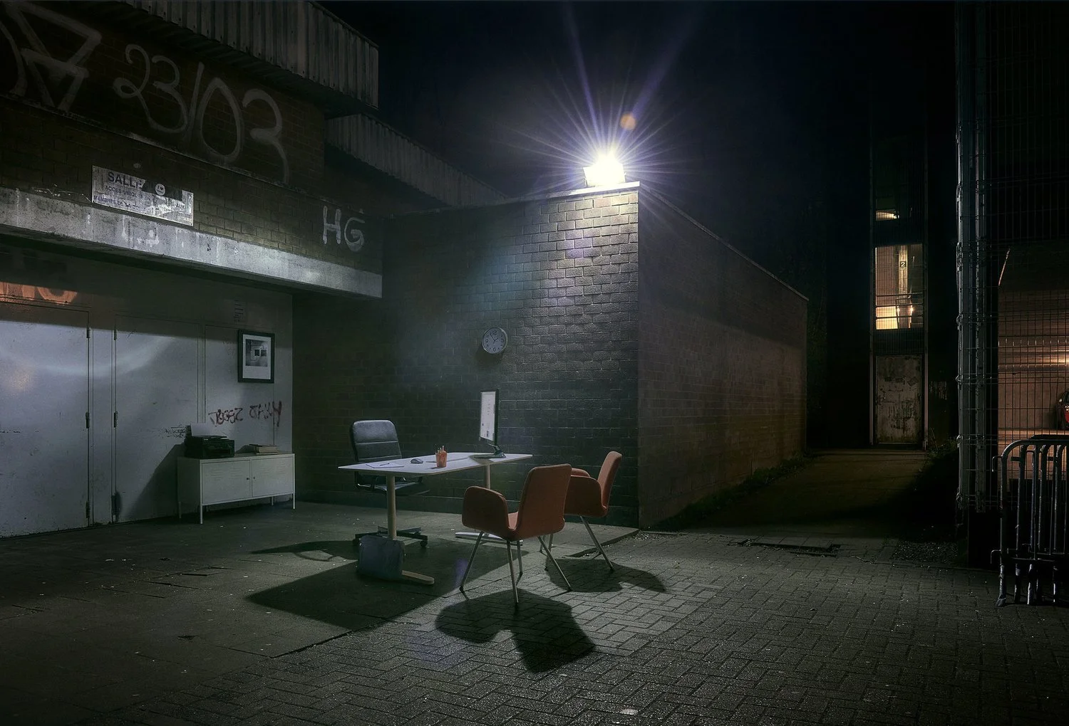 An outdoor scene at night featuring a small table with two chairs and a computer monitor, illuminated by a bright light on a brick wall, with graffiti, a clock, and a metal cabinet in the background.