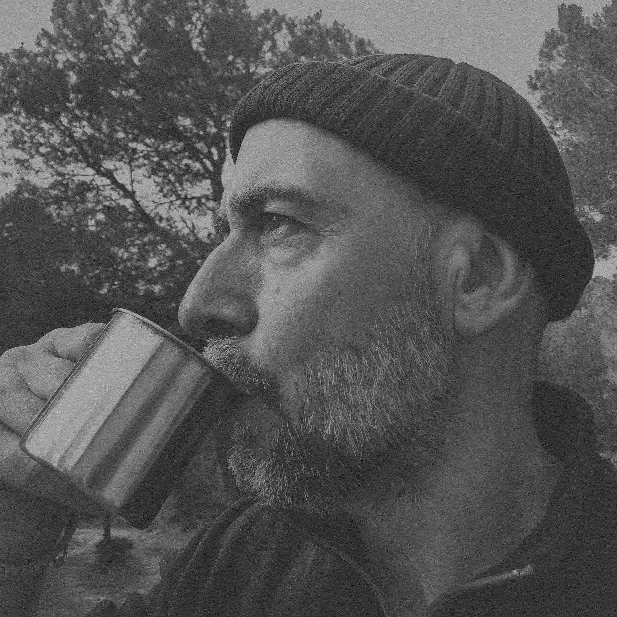 Black and white photo of a man with gray beard and mustache, wearing a knit cap, drinking from a can outdoors with trees in the background.